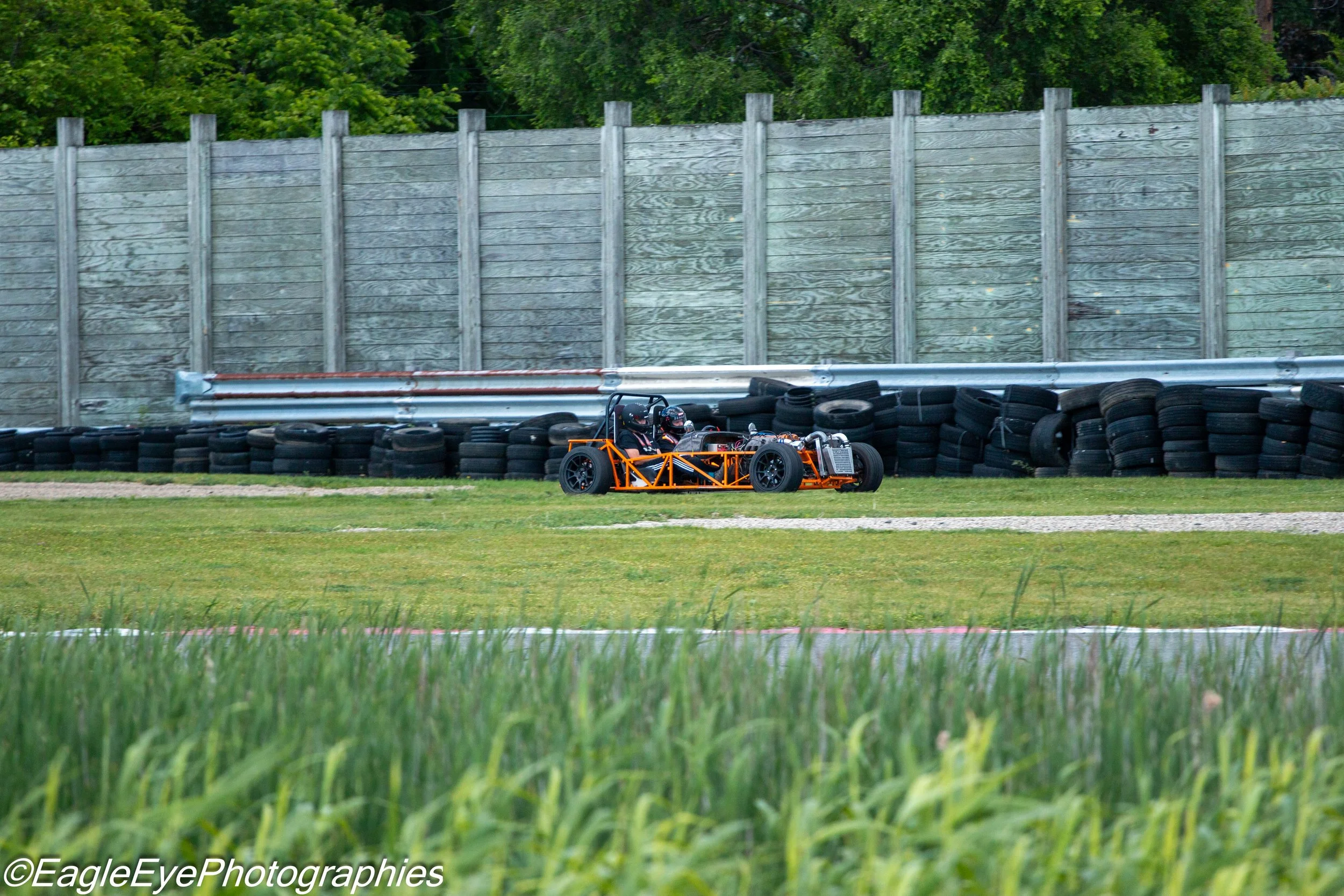 A race car on a track, with a high wooden fence and tire barriers as safety measures, surrounded by green grass and trees.