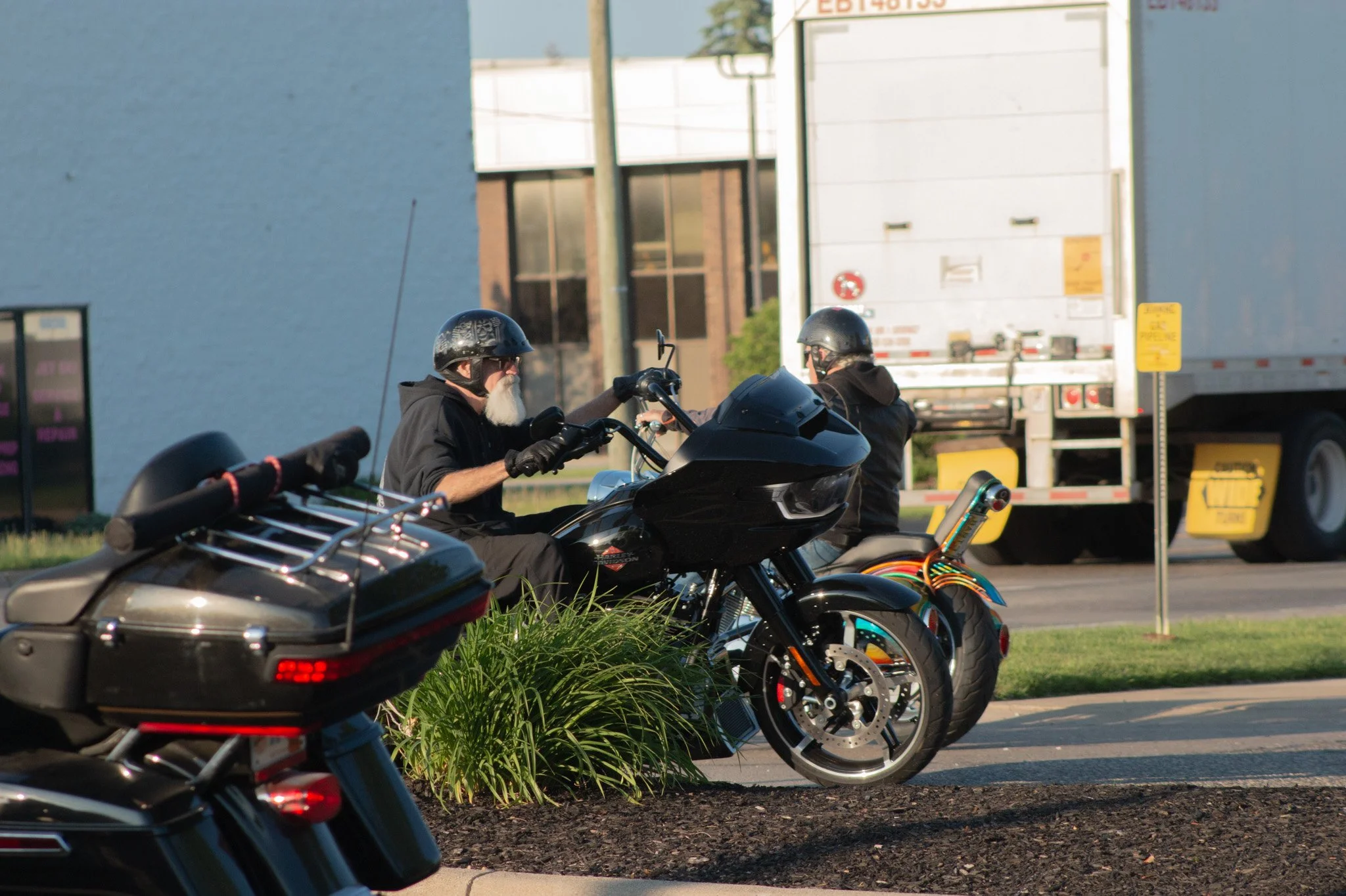 Two motorcyclists wearing helmets and black clothing riding motorcycles along a city street during daylight, with a large white truck in the background.