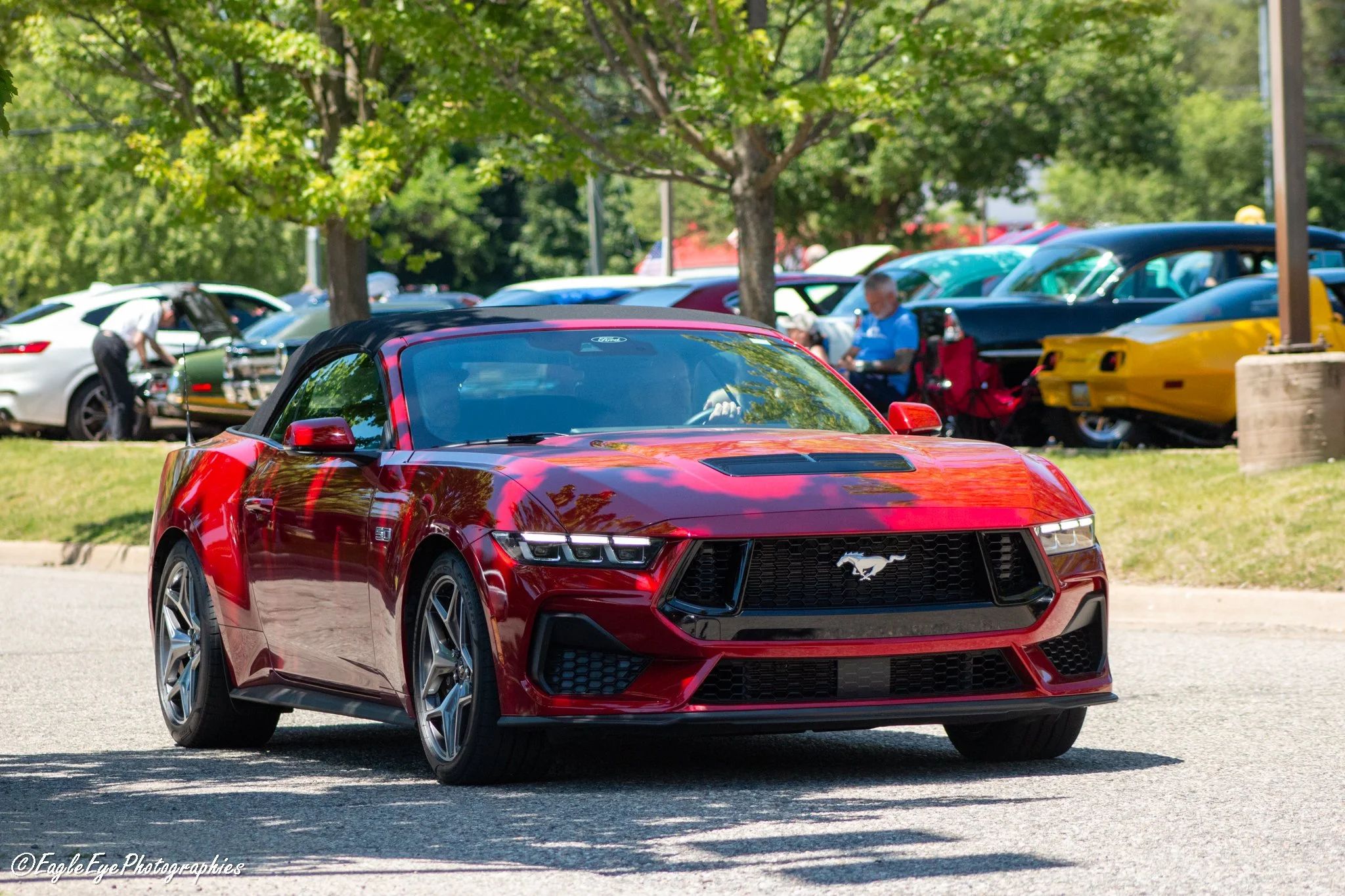 Red Ford Mustang sports car parked outdoors with other cars and people in the background, surrounded by green trees.