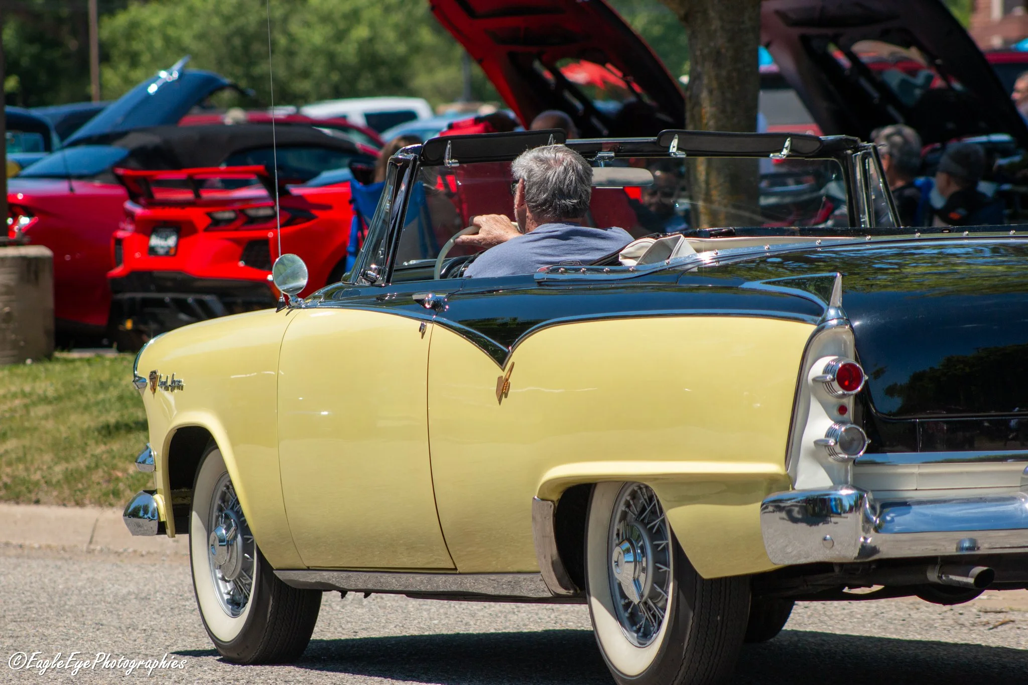 A vintage yellow convertible car with a black roof, driven by an older man with gray hair, at an outdoor car show. Several modern red and black cars are parked behind him, with their hoods open. Trees and people are visible in the background.