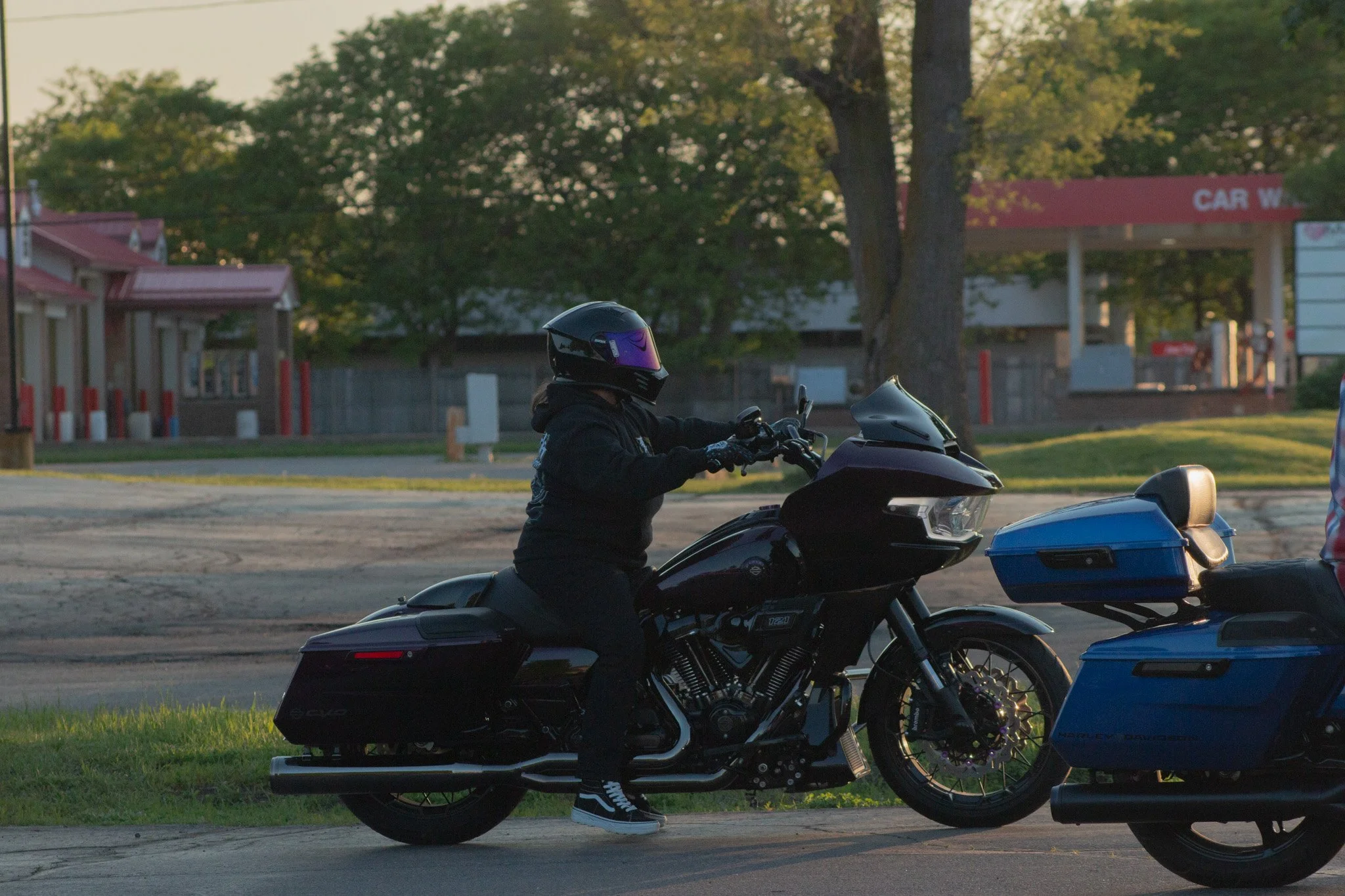 A person dressed in dark clothing and wearing a helmet is riding a black and blue motorcycle on a paved road with green grass on the side, during a sunset or sunrise. There are trees and a building in the background.