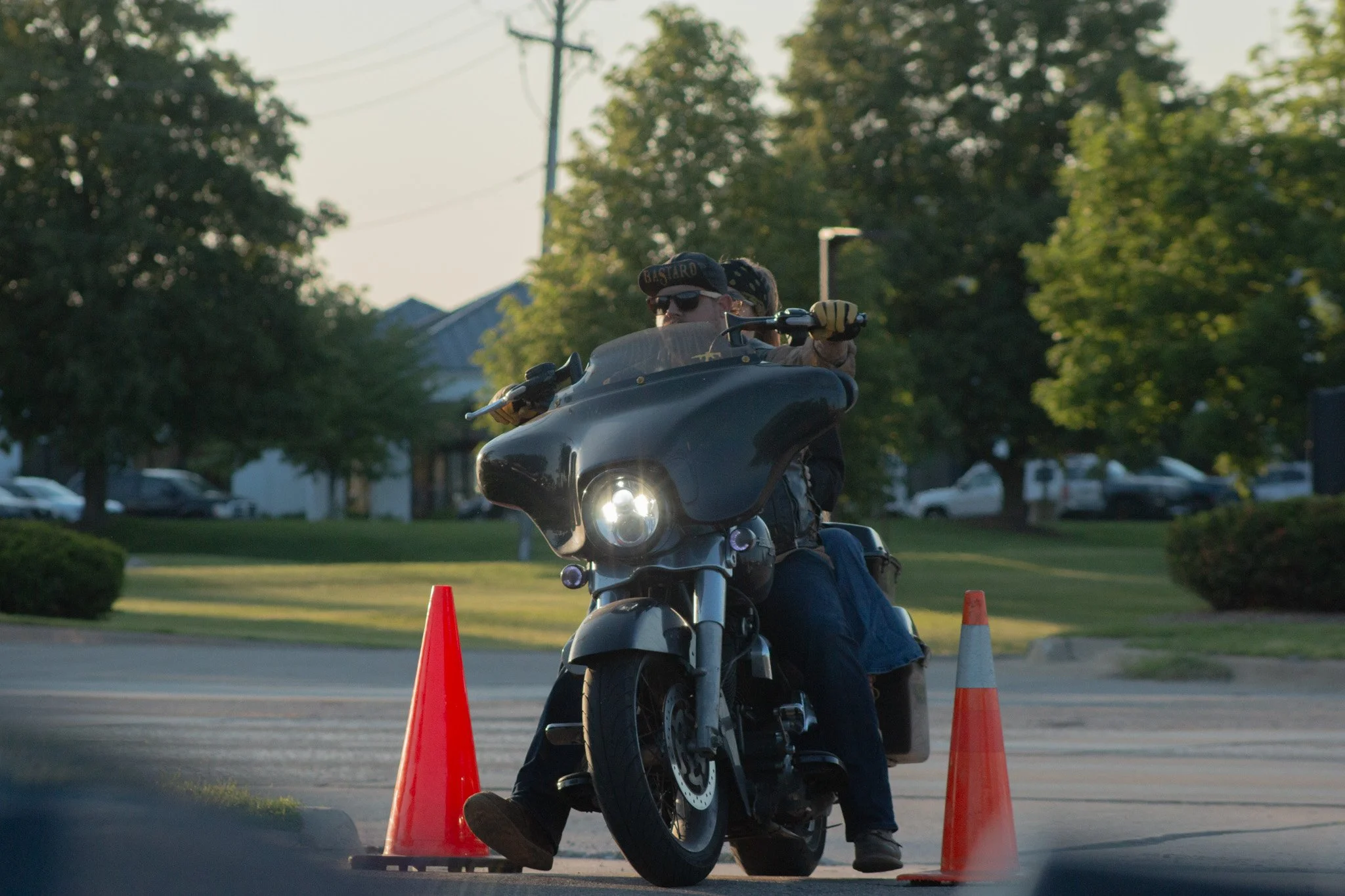 A person riding a black motorcycle between orange traffic cones in an outdoor parking lot during daytime with trees and parked cars in the background.
