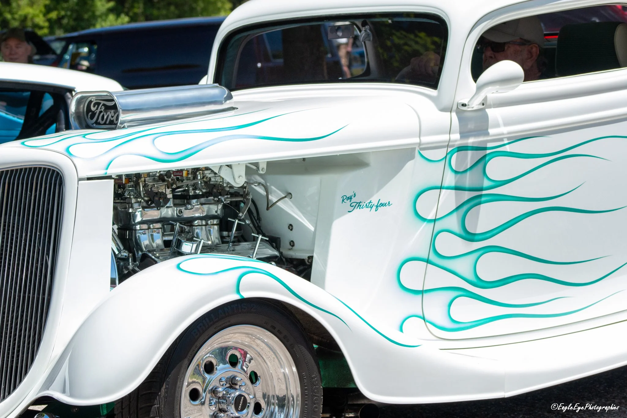 Close-up of a vintage white Ford hot rod car with teal flame decals. The car's hood is open, exposing a chrome engine. The car has a shiny chrome wheel and a black tire. Reflections of people and trees are visible on the car's windows and body.