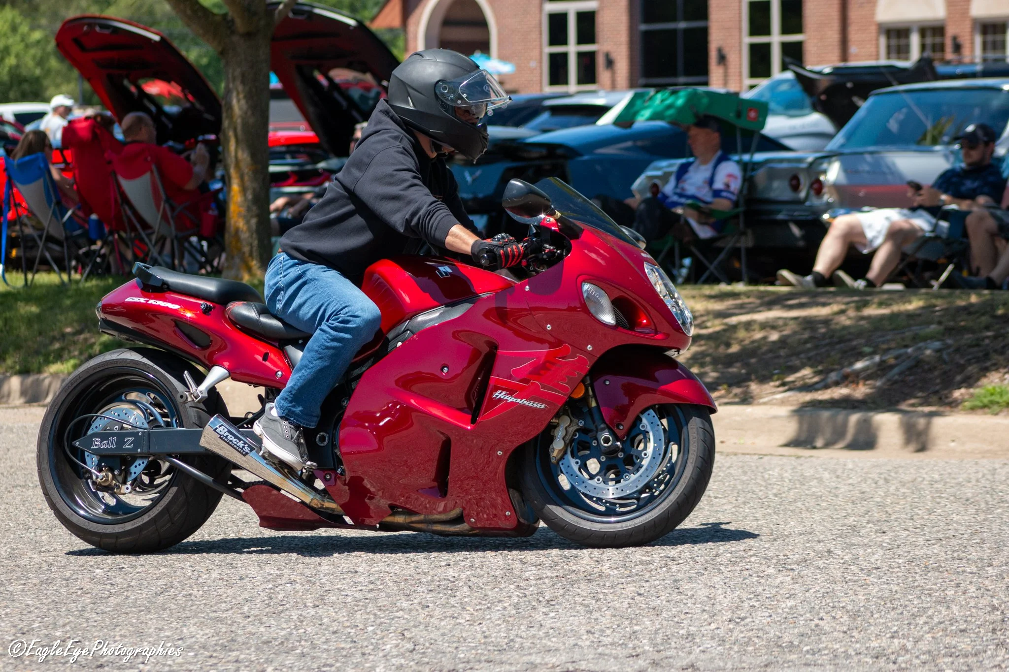 A person wearing a black helmet riding a red motorcycle on a street. There are people sitting and standing under umbrellas in the background.