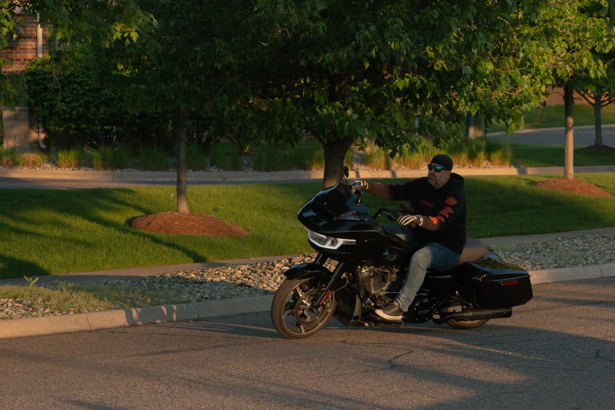 A man riding a black motorcycle on a suburban street during sunset, wearing sunglasses, a black jacket, and gloves, with trees and green grass in the background.