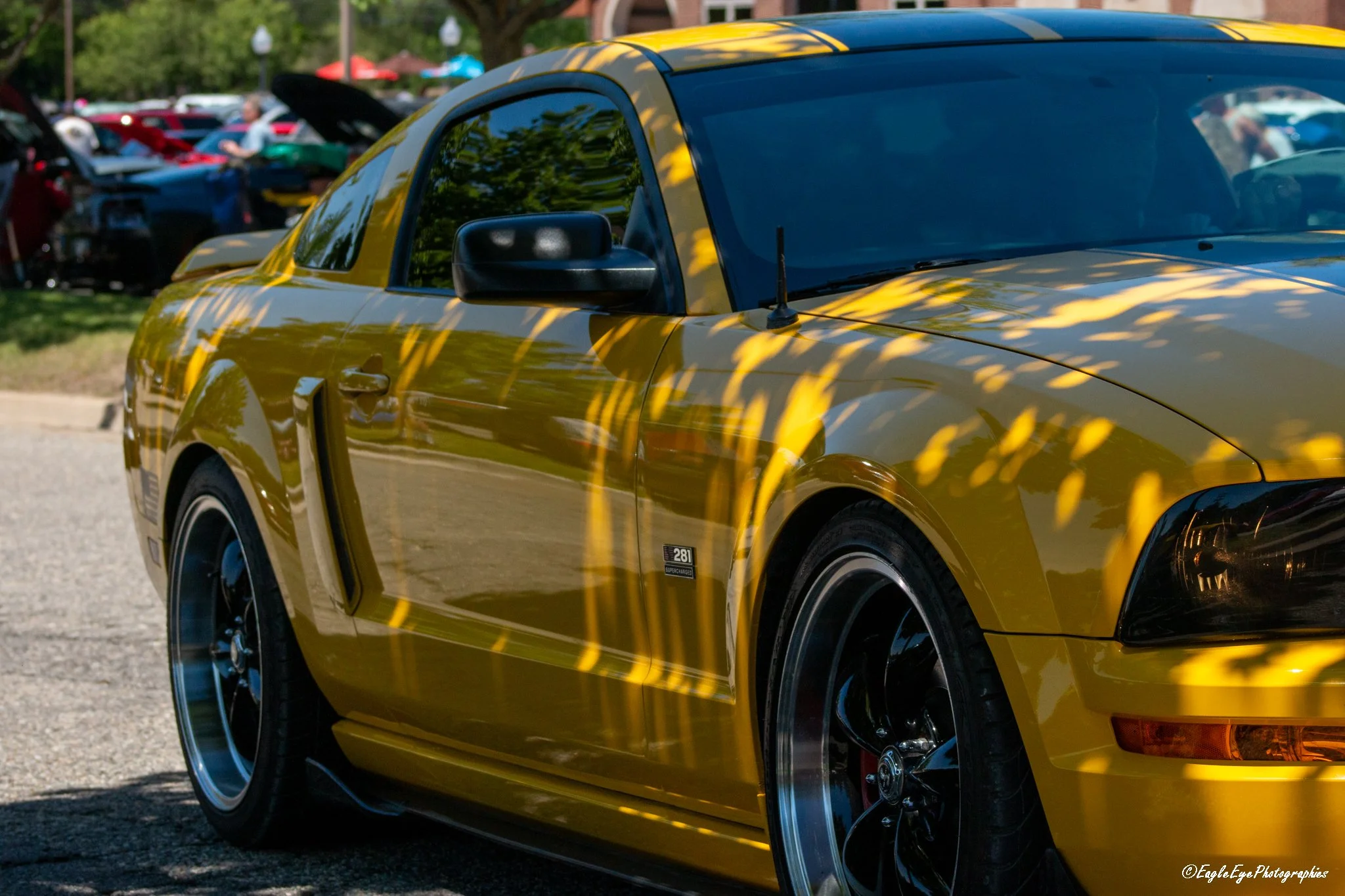 Close-up of a yellow sports car parked at an outdoor event with other cars and people in the background, reflected in the car's surface.