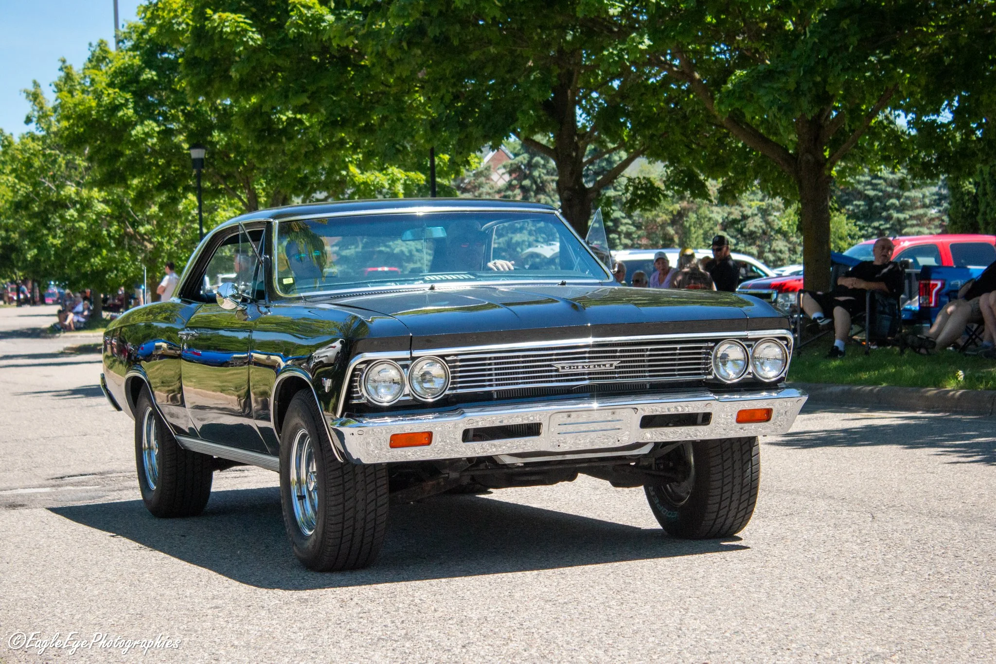 A vintage black Chevrolet Chevelle muscle car parked outdoors under green leafy trees at a car show, with people in the background.