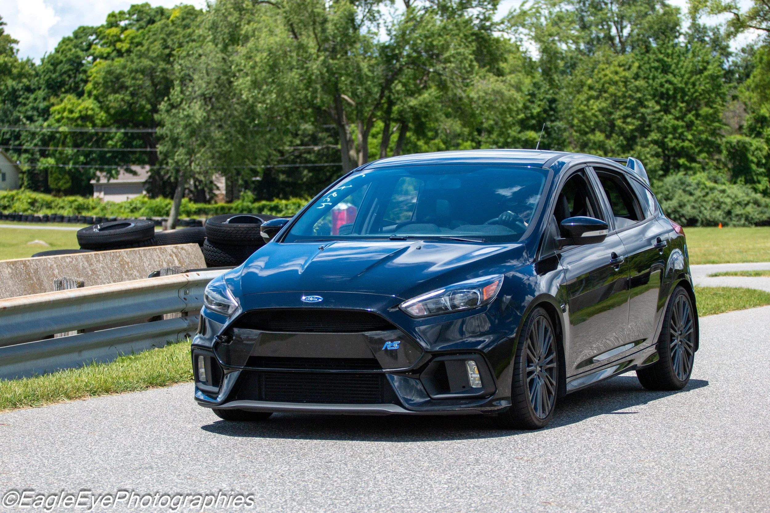 A blue Ford Mustang Mach-E electric SUV parked by a metal guardrail on a paved roadside, with stacks of tires and a concrete barrier nearby, surrounded by lush green trees and a partly cloudy sky.