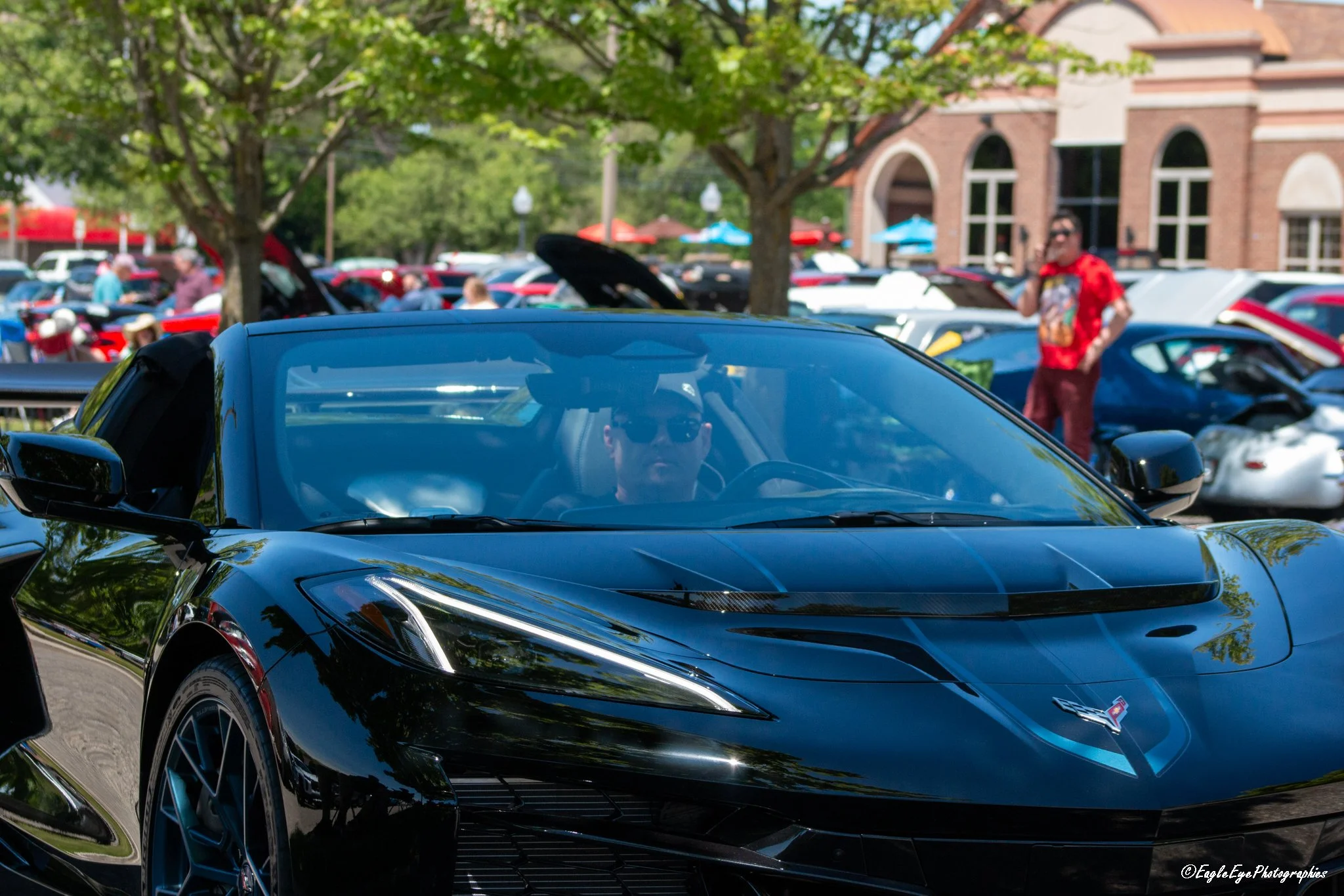A black sports car is parked outdoors at a car show, with a person visible through the windshield. In the background, there are many other cars, people, trees, and a building with a brick facade and arched windows.