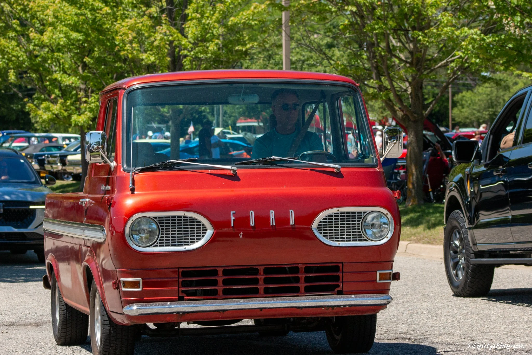 A vintage red Ford van parked among other vehicles at an outdoor car show with trees in the background.
