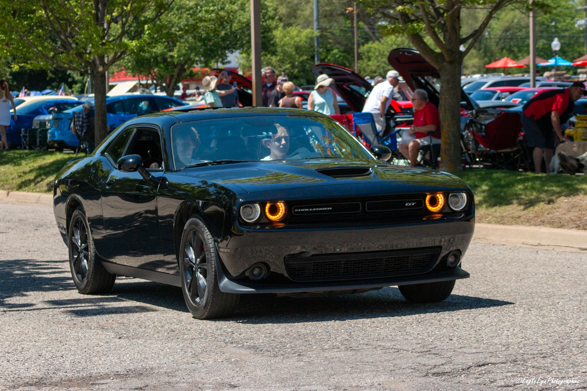 Black Dodge Challenger GT with illuminated orange headlights driving past a crowded outdoor car show with various cars and people under trees.