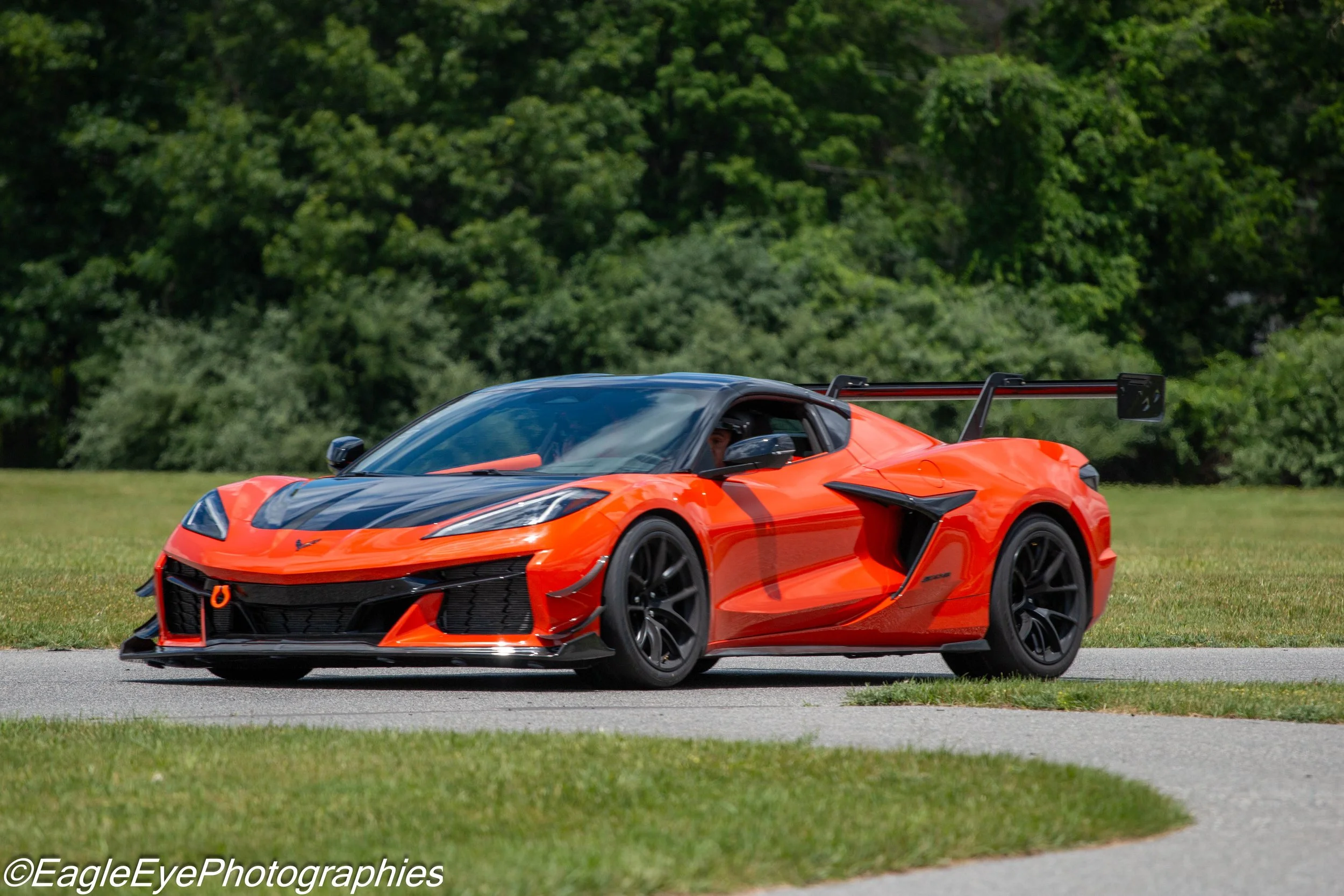 An orange sports car with a black roof and spoiler driving on a paved road with grass and trees in the background.