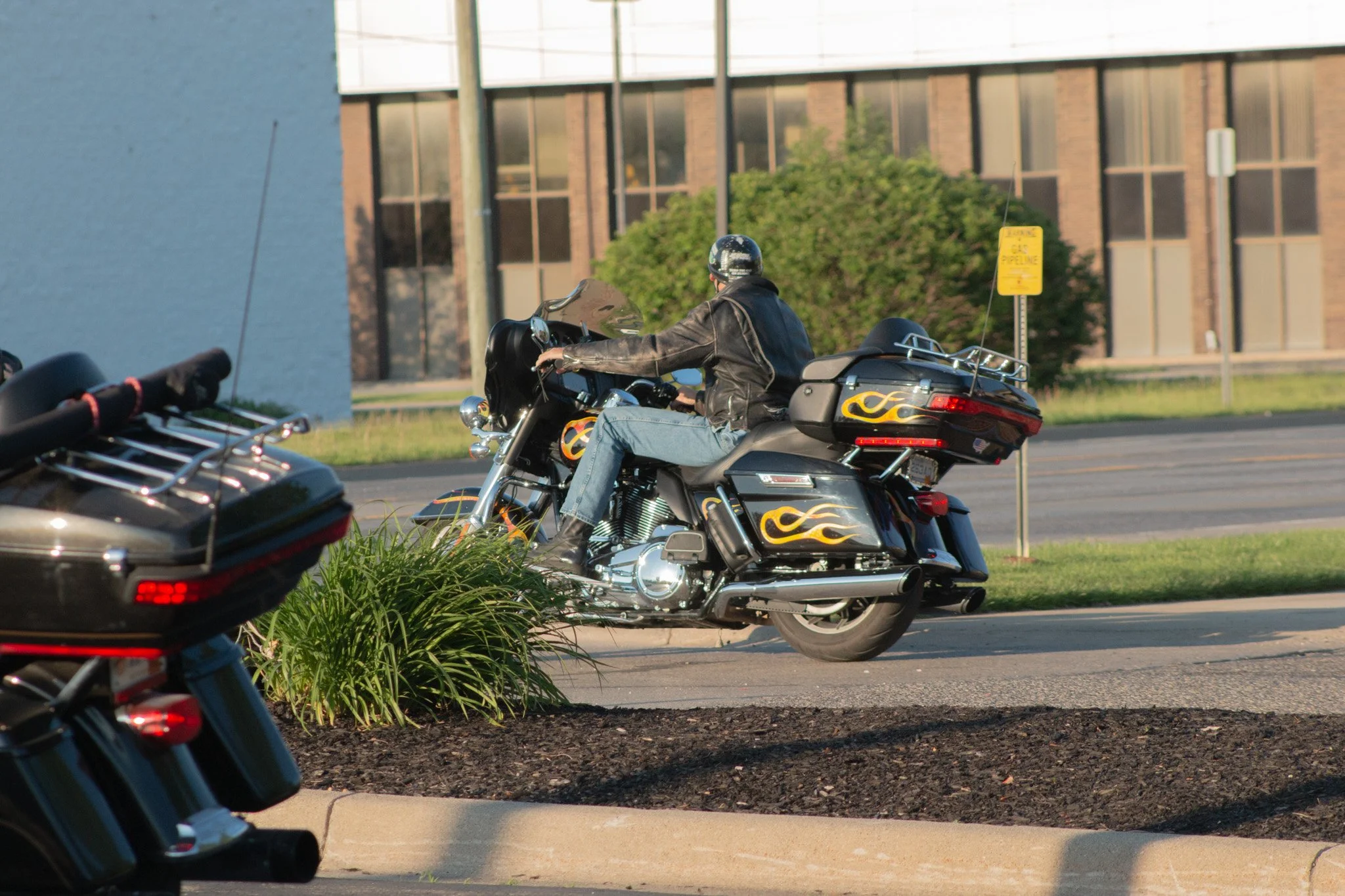 A man riding a motorcycle with flames painted on the side, wearing a leather jacket and helmet, is riding on a city street near a brick building and a yellow pipeline sign. Another motorcycle is partially visible in the foreground.