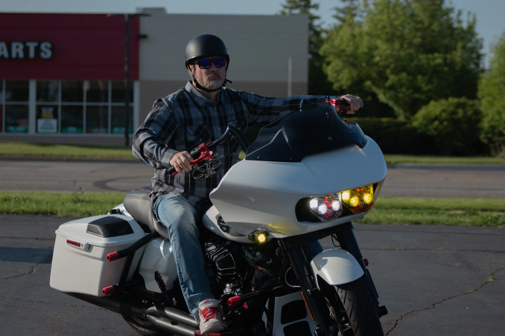 A man wearing sunglasses and a helmet riding a white touring motorcycle with storage compartments, parked on a paved surface outside a commercial building with a red sign and green trees in the background.