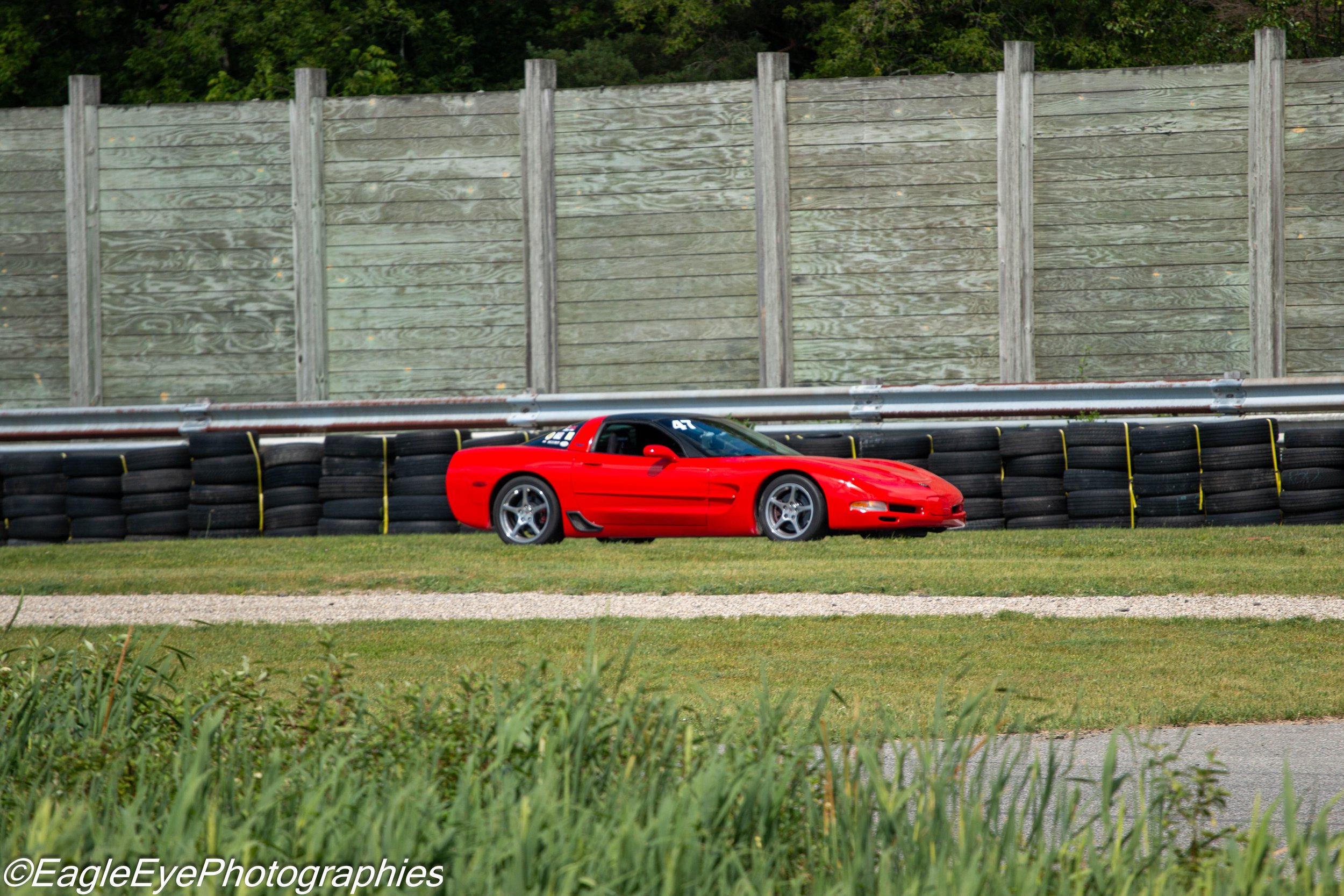A red sports car on a racetrack with a tall wooden sound barrier and green trees in the background.