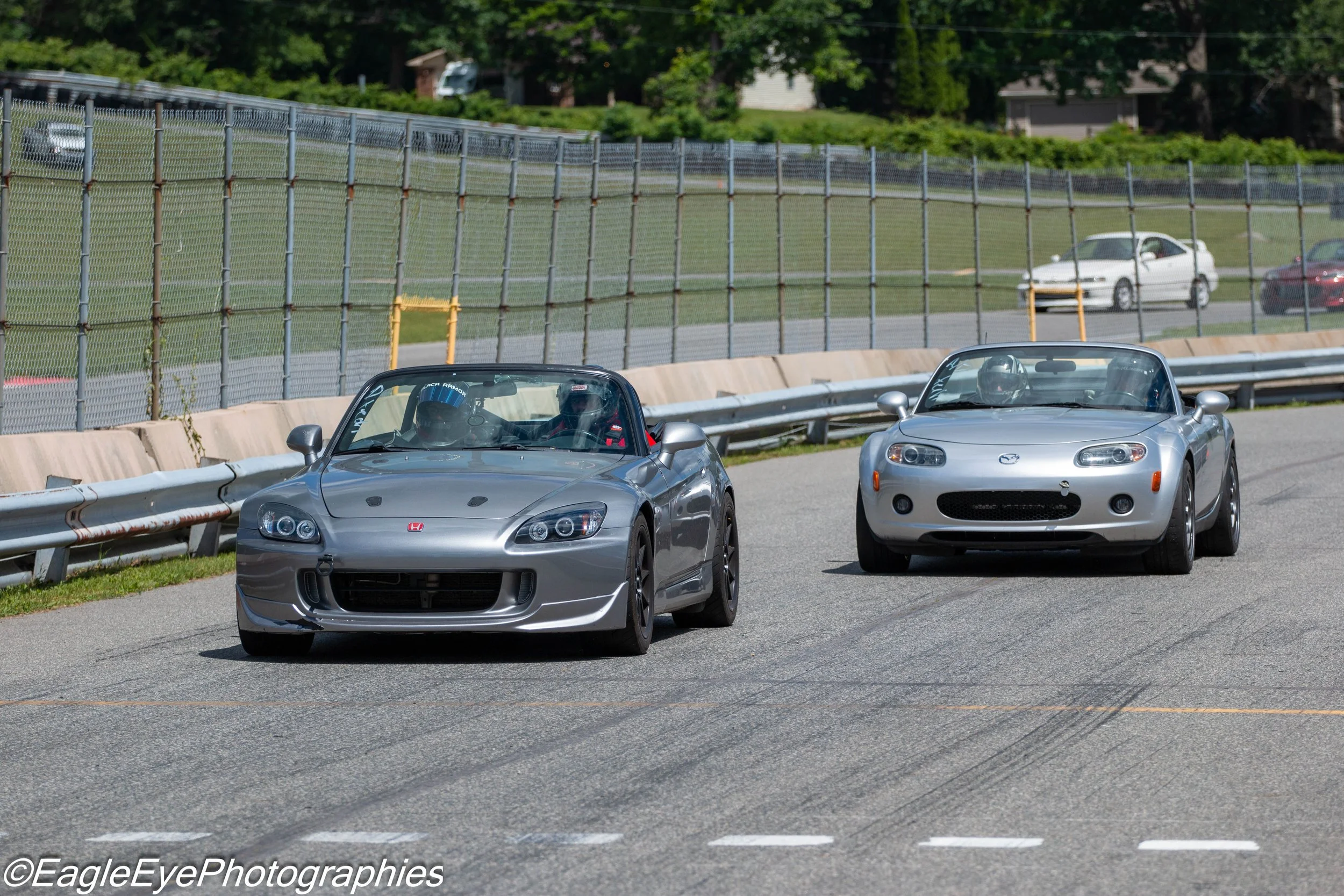 Two silver Mazda MX-5 Miata sports cars on a racetrack, with drivers wearing helmets inside, surrounded by safety barriers and a tall fence, and greenery in the background.
