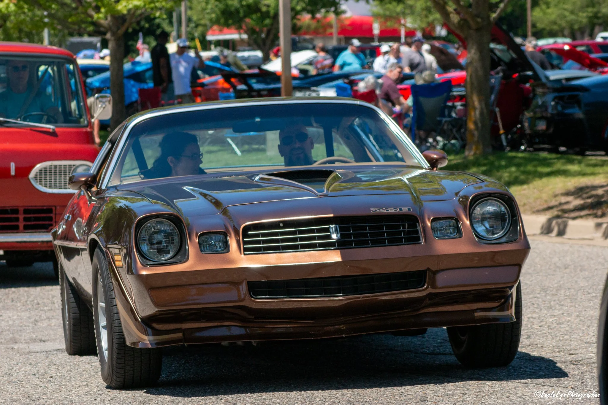 A bronze-colored vintage Chevrolet Camaro Z28 driving at a car show with people and other classic cars in the background.