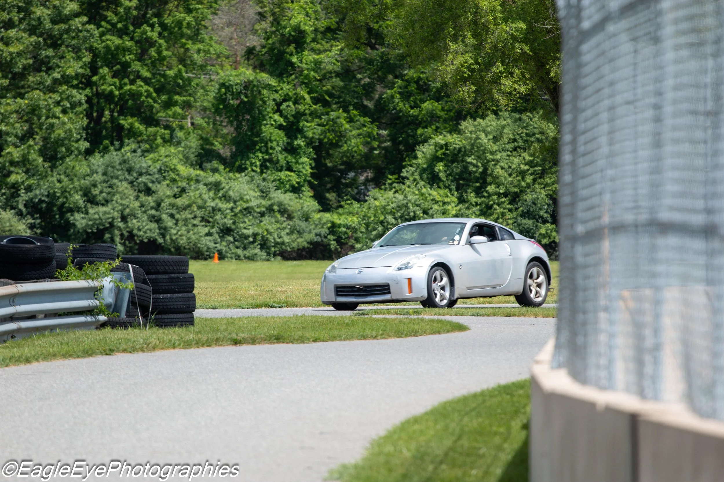A silver sports car parked on a grassy area near a row of stacked tires and a barrier with green trees in the background.