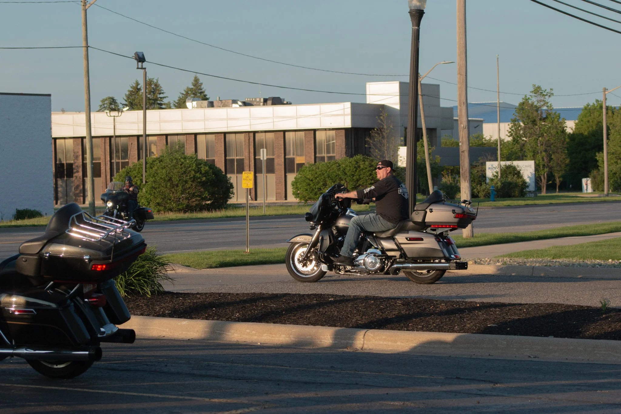A man riding a black and gray motorcycle on the street, with another motorcycle parked nearby. The scene is set in a suburban area with trees, power poles, and building structures in the background during the daytime.