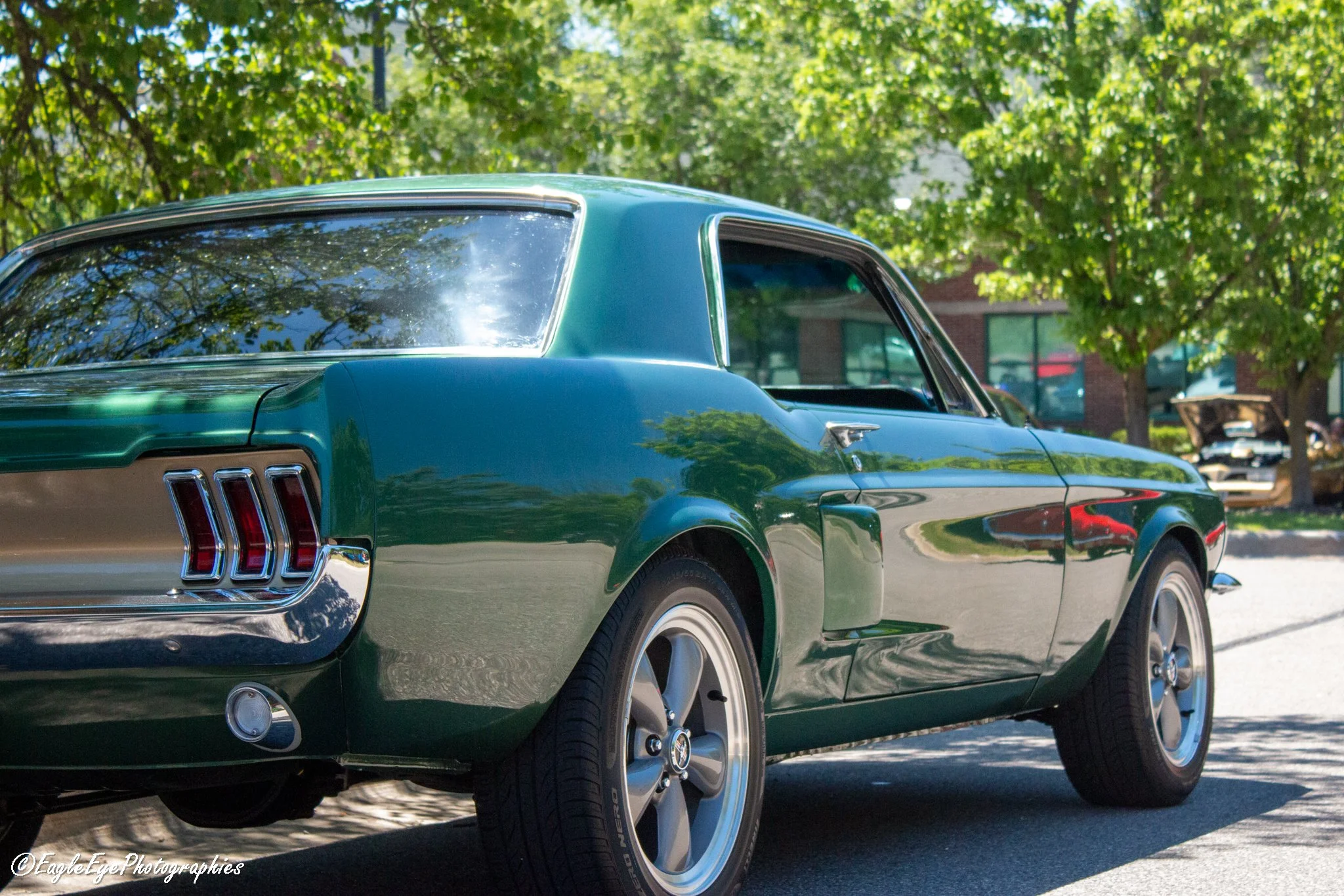 A vintage dark green Ford Mustang car parked under trees with a building and other vehicles in the background.