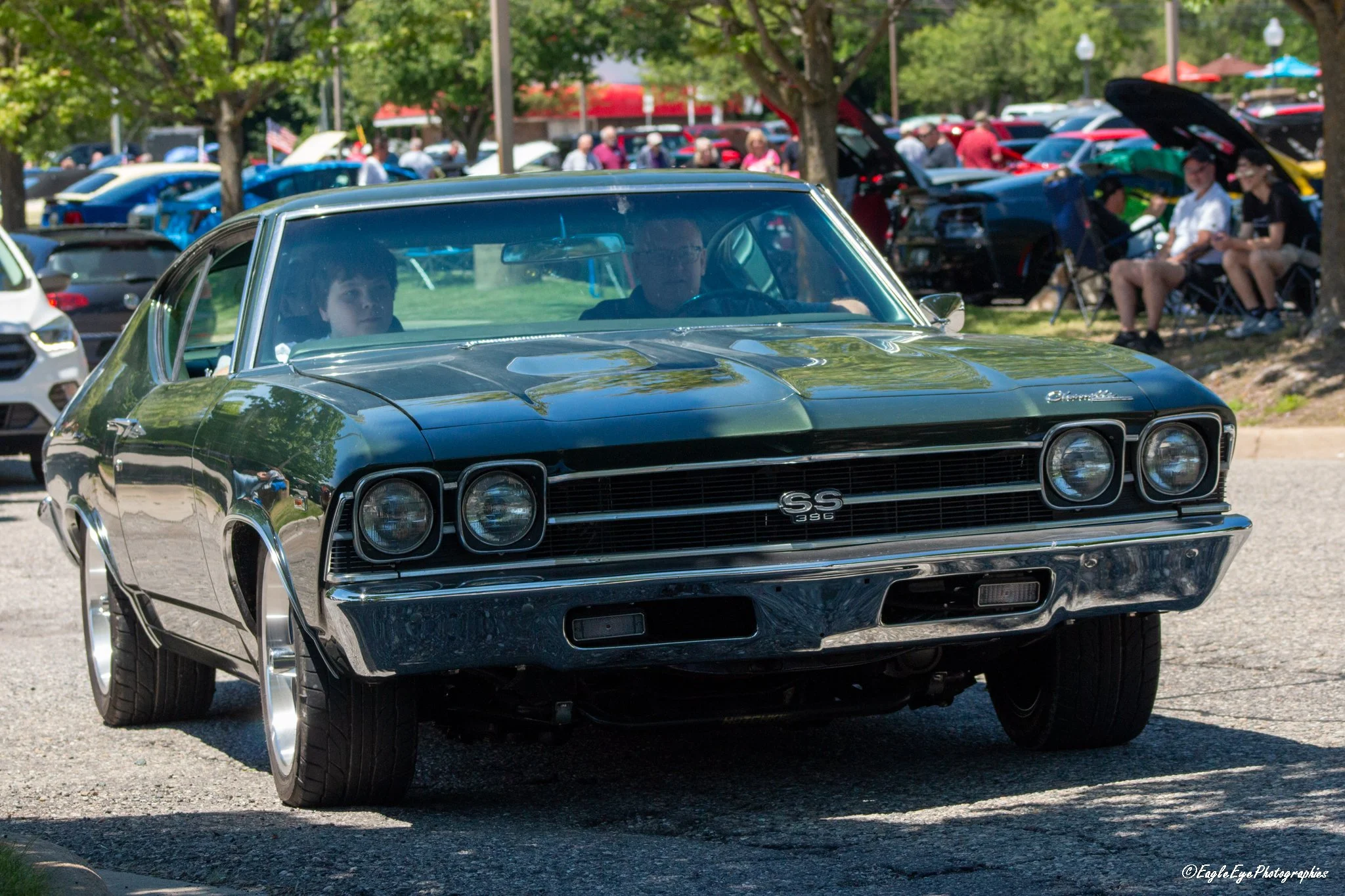 A black vintage Chevrolet SS396 muscle car parked at an outdoor car show with people walking and sitting in the background.