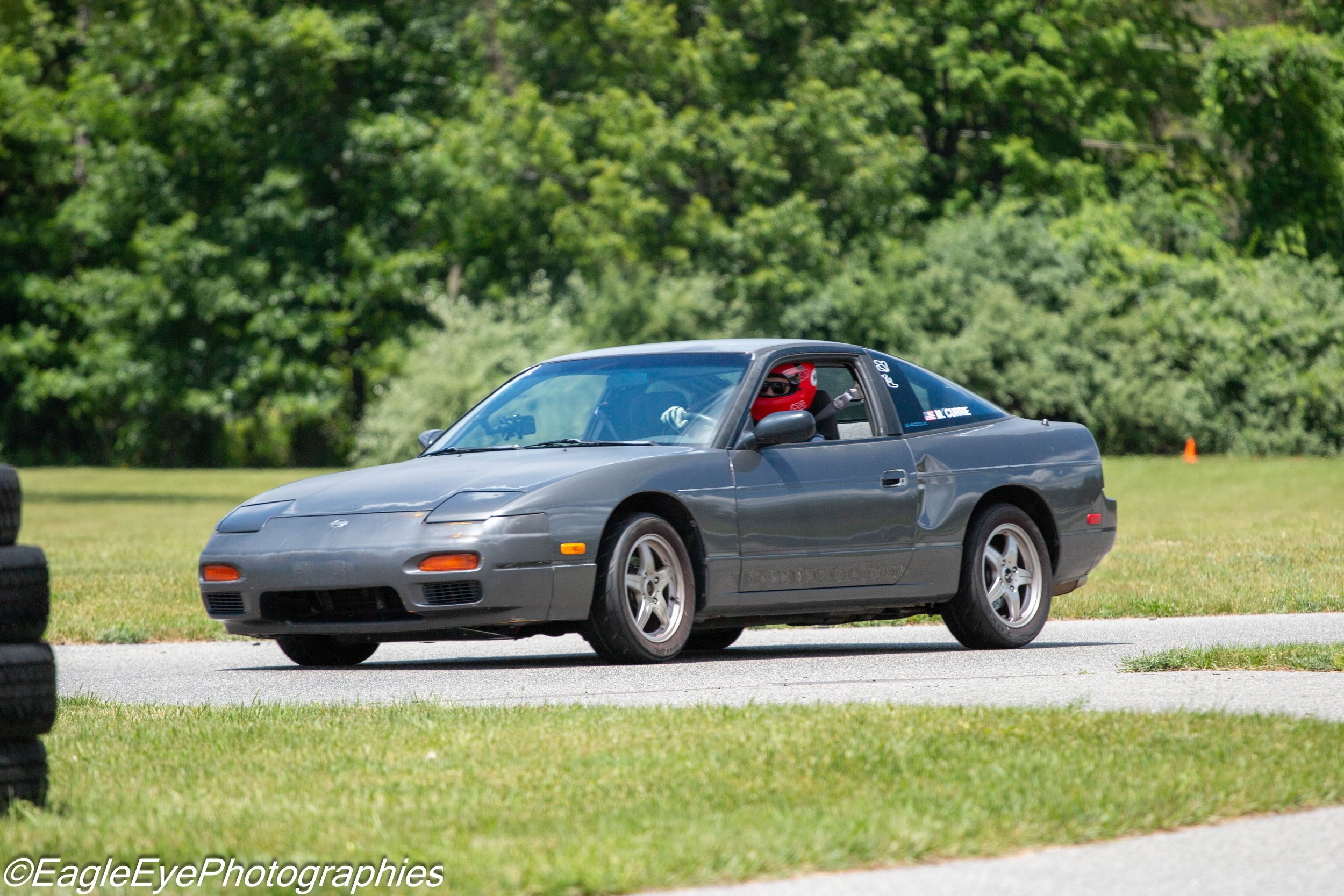 A gray sports car with a person wearing a red helmet driving on a grassy area during a car racing event, with trees in the background.
