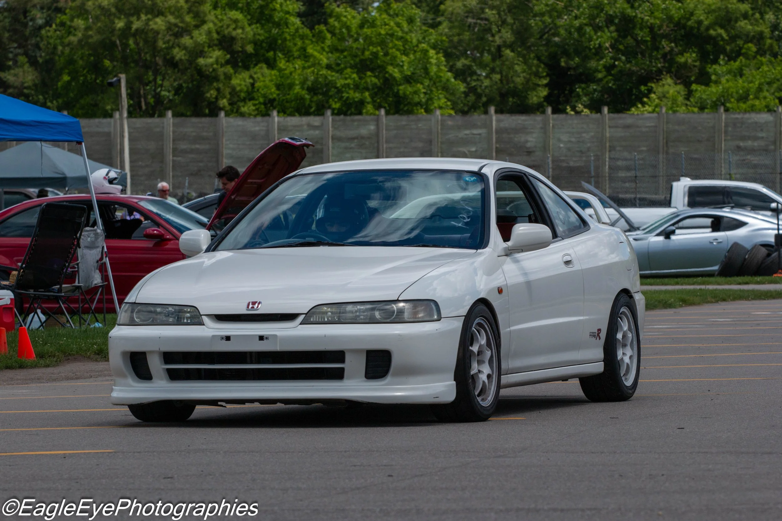 A white Honda sports car driving on a parking lot with yellow lines, with other cars and a person in the background. The scene suggests a car event or meet.