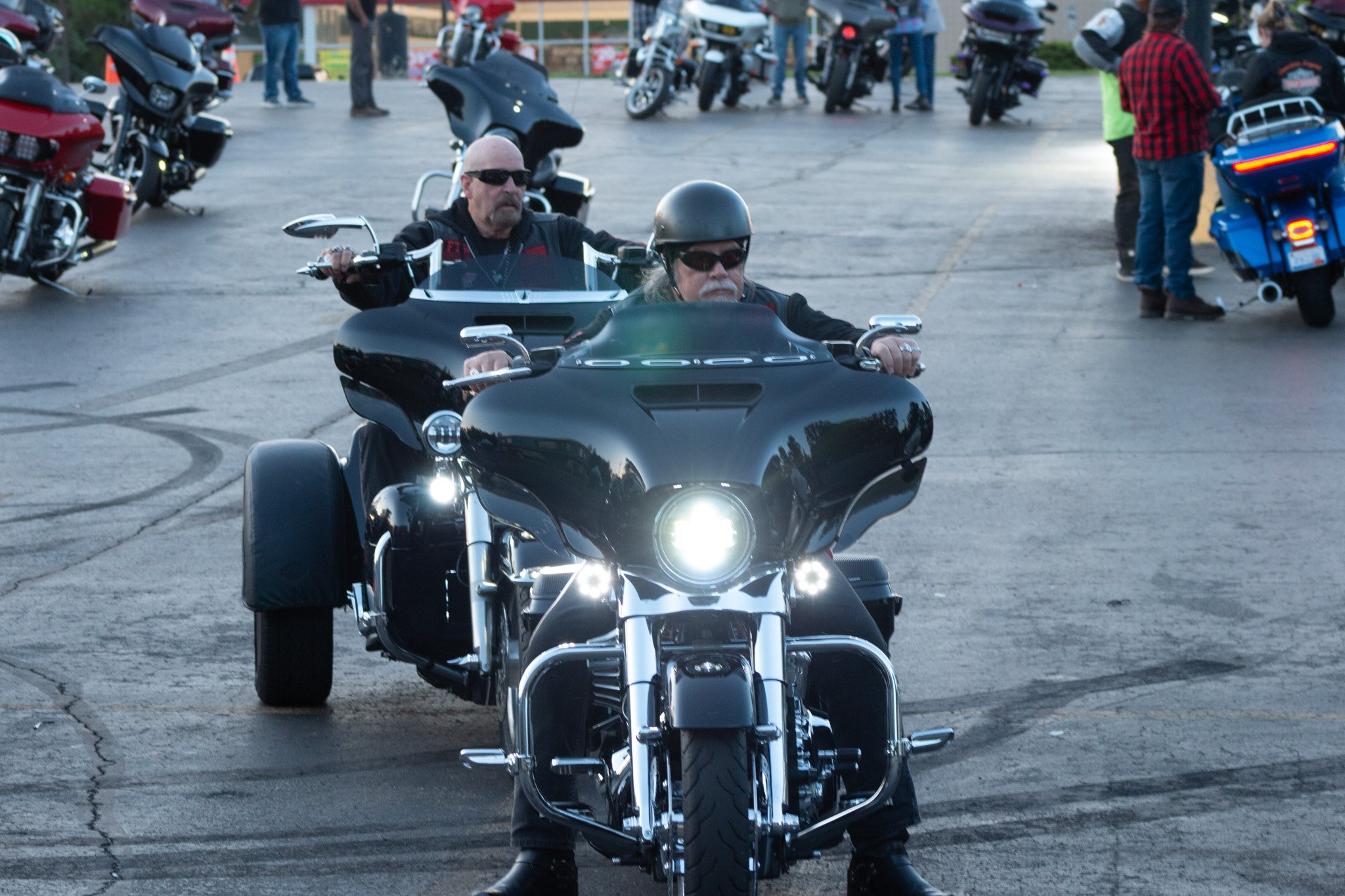 Two men riding black motorcycles, wearing helmets and sunglasses, at a motorcycle gathering in a parking lot with other motorcycles and people in the background.