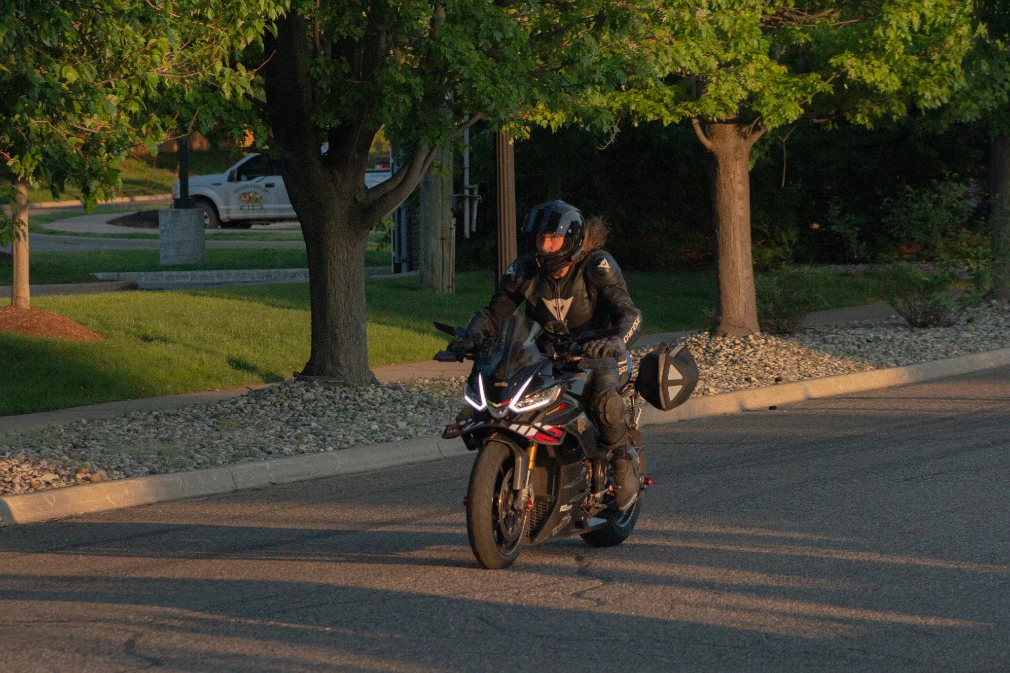 A person riding an electric motorcycle on a residential street during dusk, with trees, a park, and a white vehicle in the background.