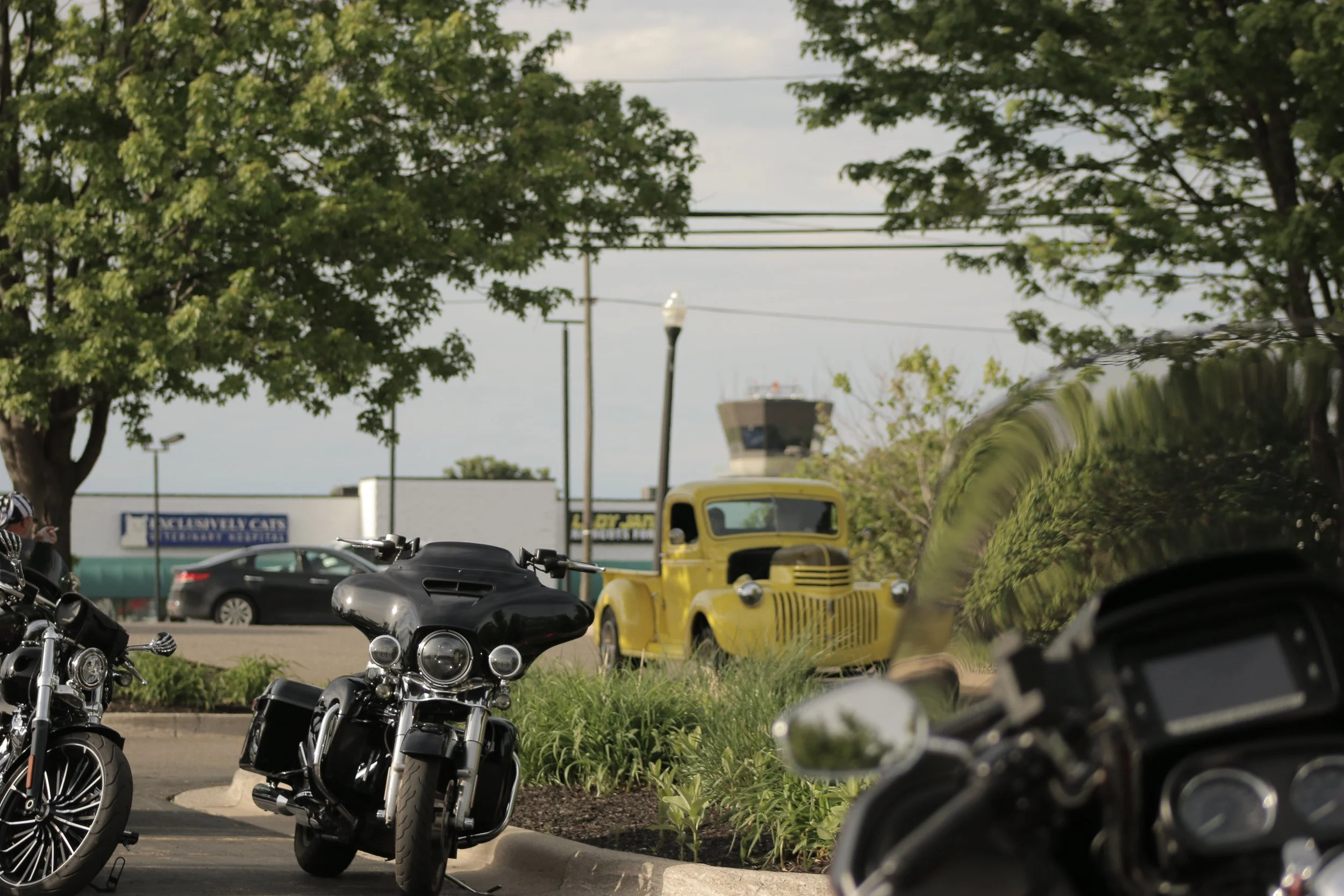 Motorcycles parked in a lot with trees, a yellow vehicle, and a control tower in the background.