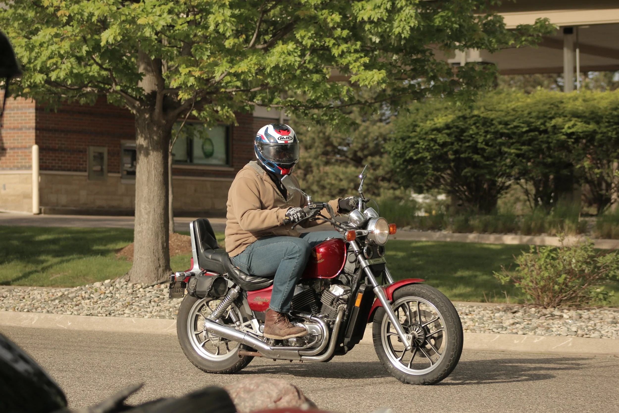 Person wearing a helmet and tan jacket riding a red motorcycle on a city street with trees and buildings in the background.