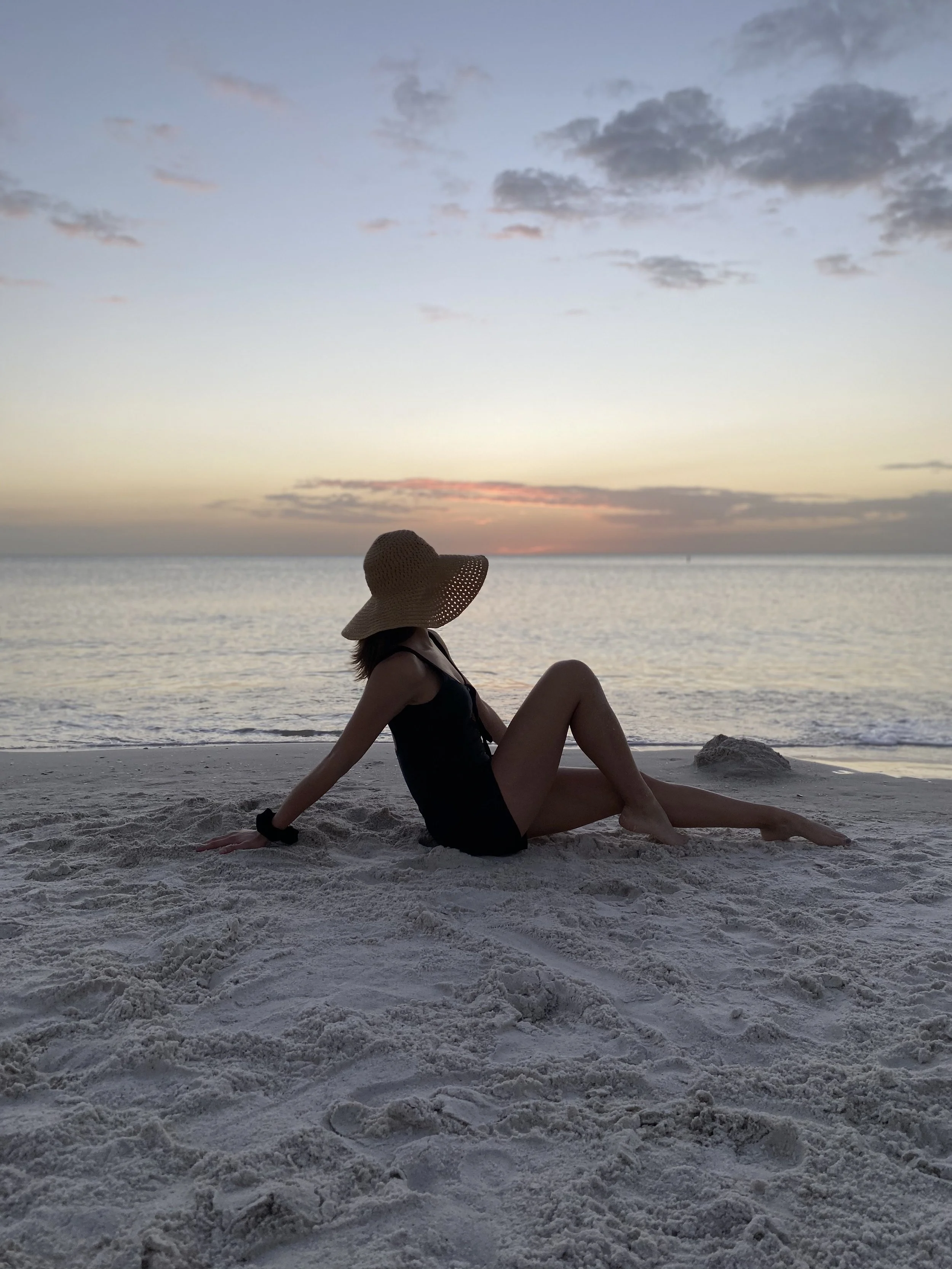 A woman wearing a straw hat and black swimsuit sitting on the sandy beach near the water during sunset, with cloudy sky above.