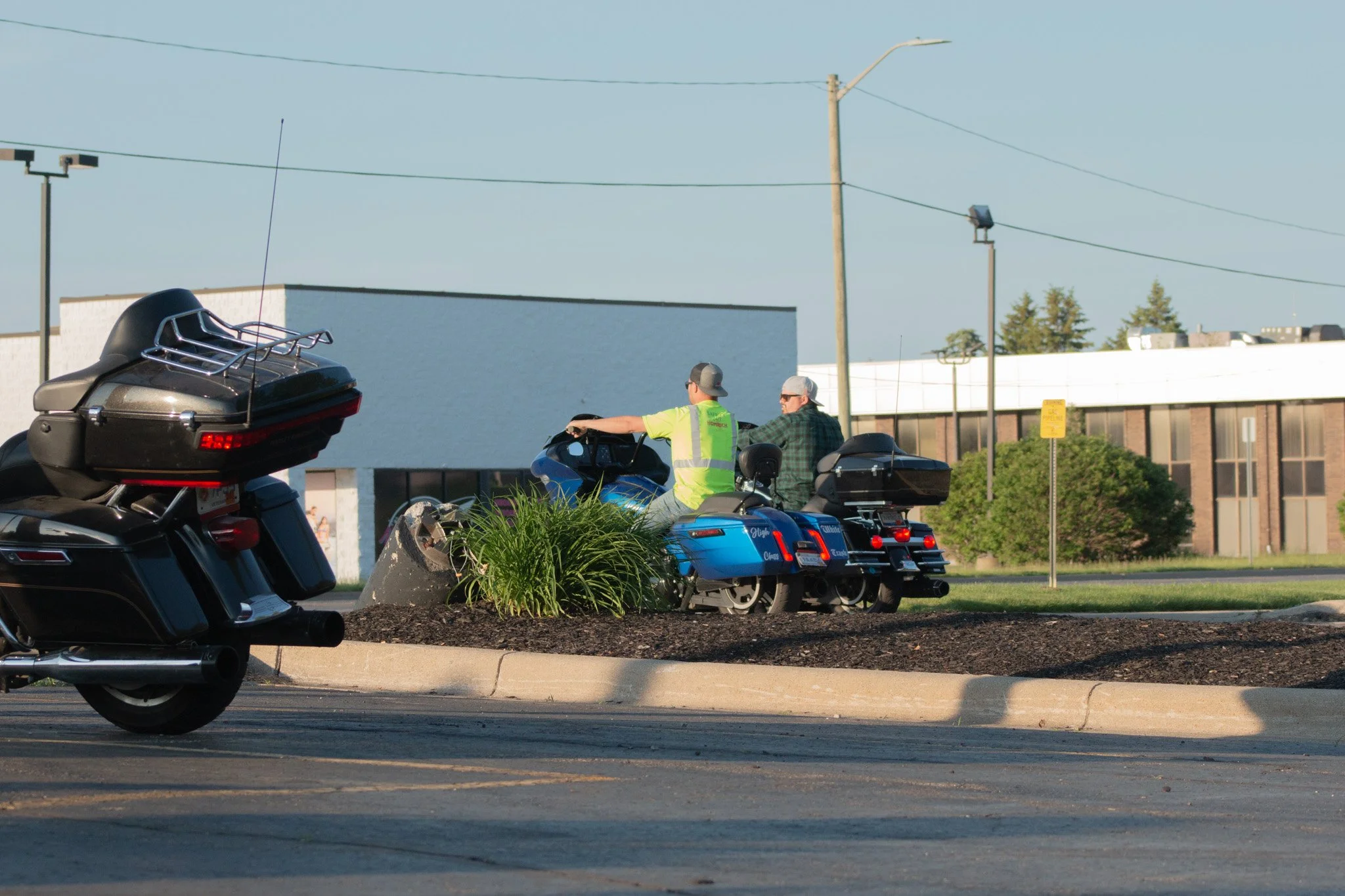 Two men riding motorcycles on a street, with a black motorcycle in the foreground and a blue motorcycle near a landscaped area with grass and shrubs. One man is wearing a neon yellow safety vest and sunglasses, and the other man is wearing a plaid sh