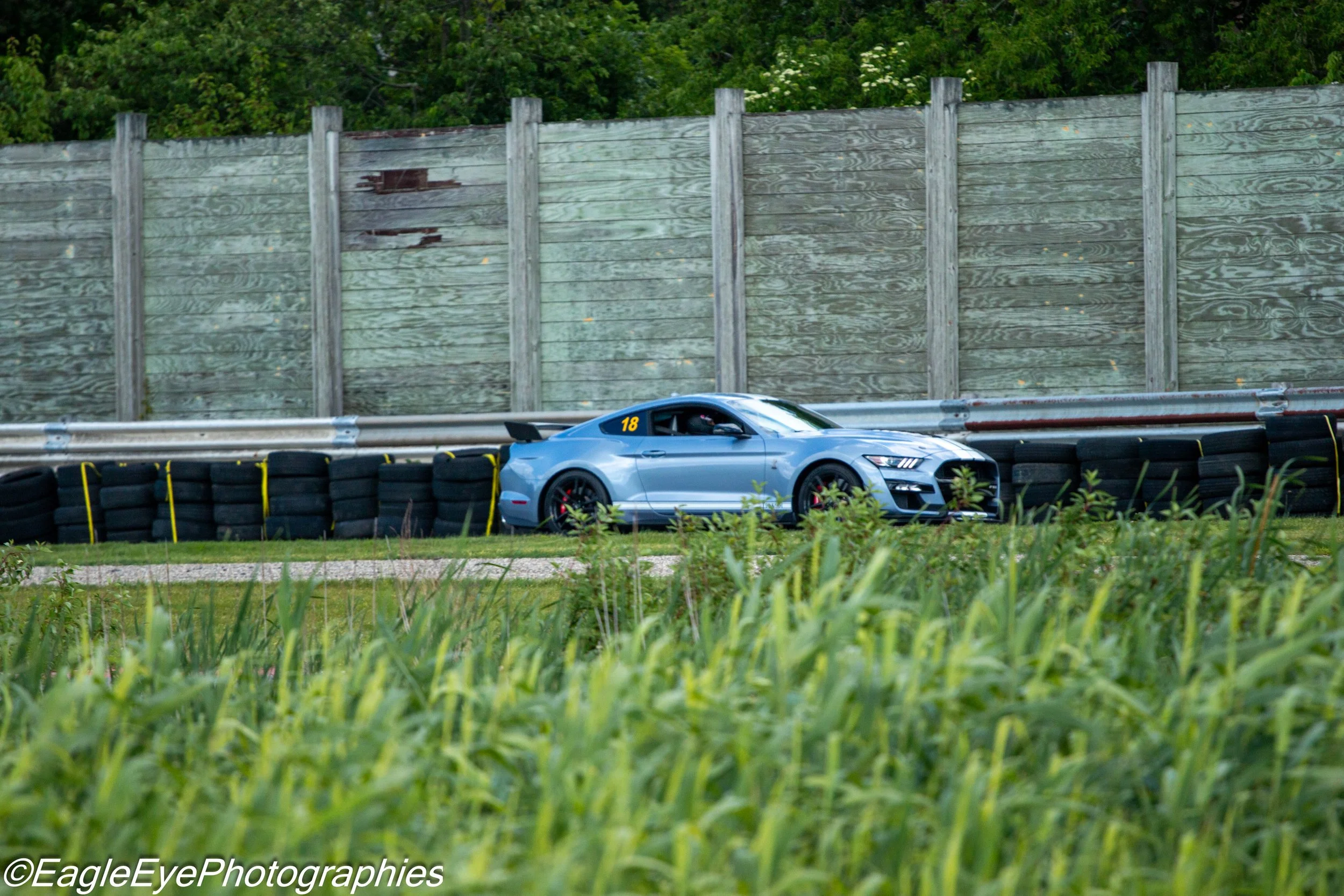 A silver sports car with the number 18 on its side is parked behind a barrier of black tires with yellow ties on an asphalt racetrack. Behind the car, there is a tall wooden fence with visible wear and some peeling paint, and behind it, there are gre