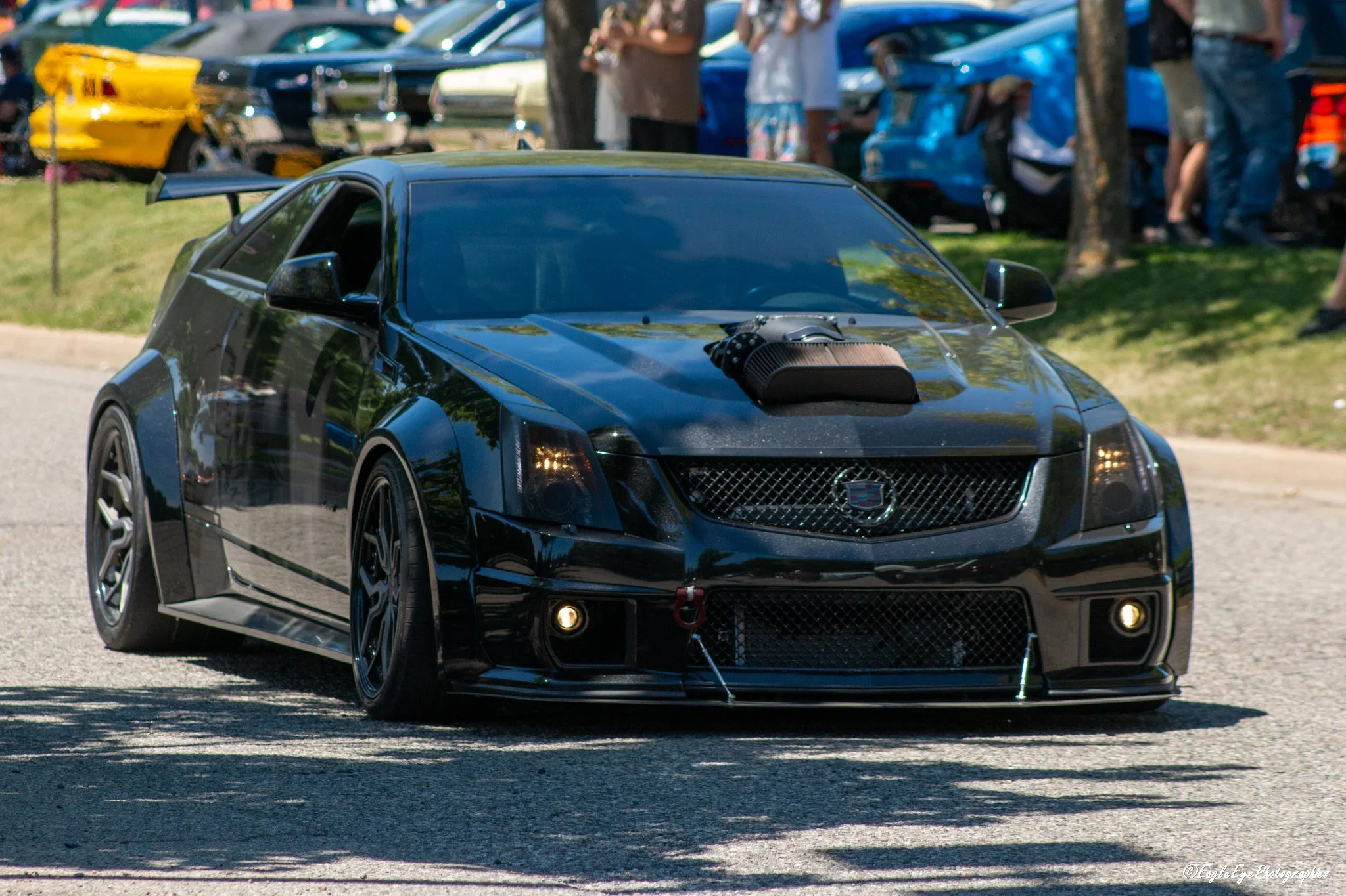 A black sports car with a large rear spoiler and aftermarket modifications, driving on a sunny street during a car show, with people and trees in the background.