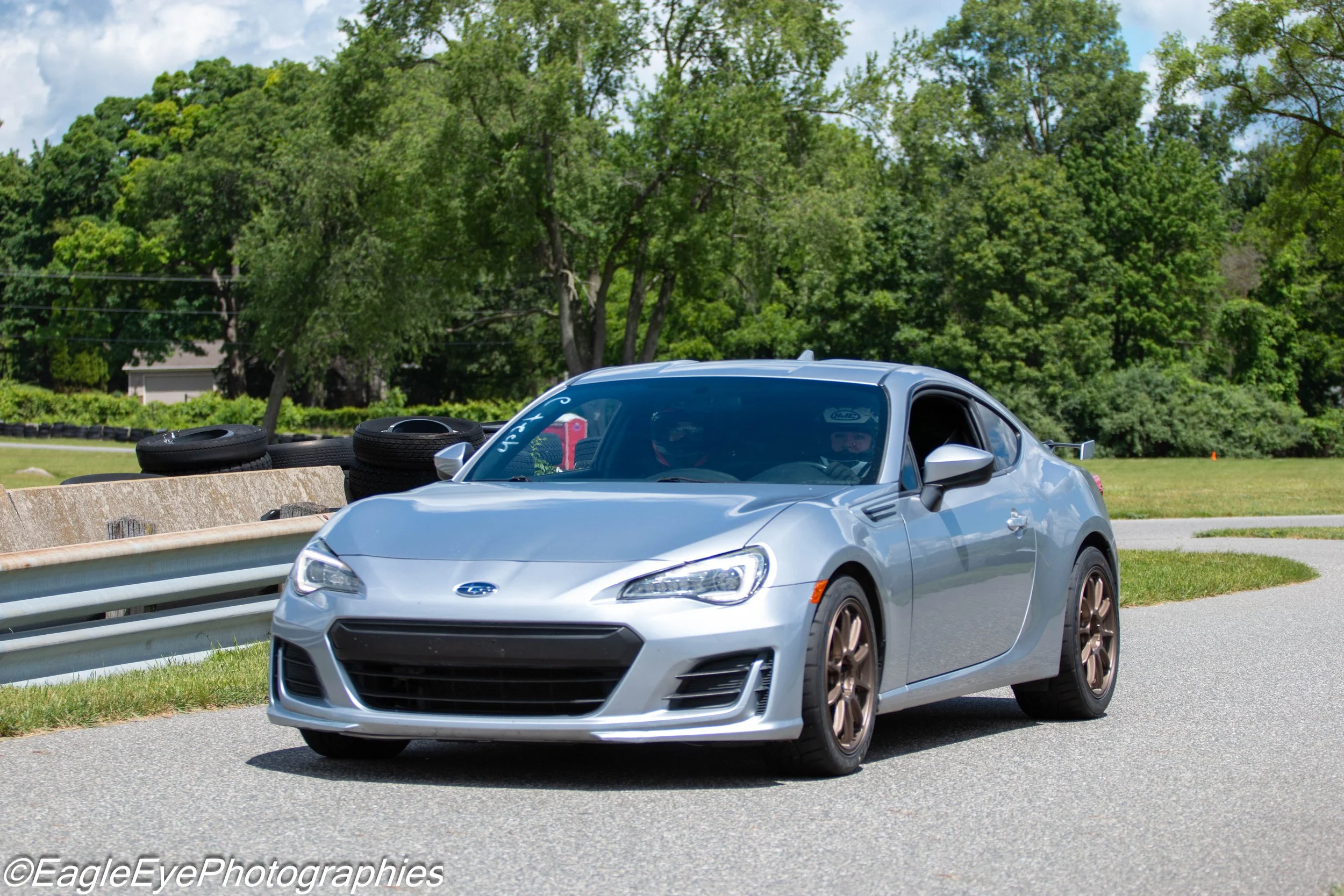 A silver sports car with bronze-colored wheels parked on the side of a street near a grassy area and trees in the background.