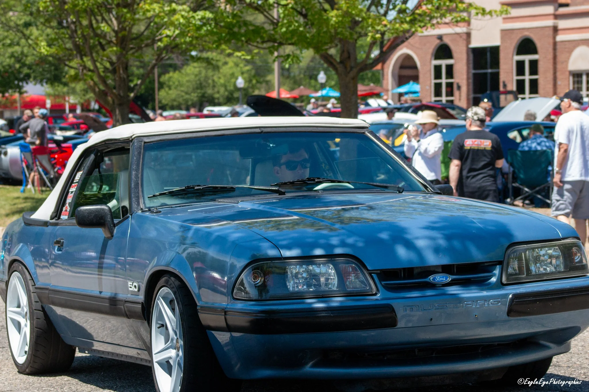A 1980s blue Ford Mustang with white wheels parked at an outdoor car show, with a crowd of people, many wearing sunglasses and hats, and tents and trees in the background.