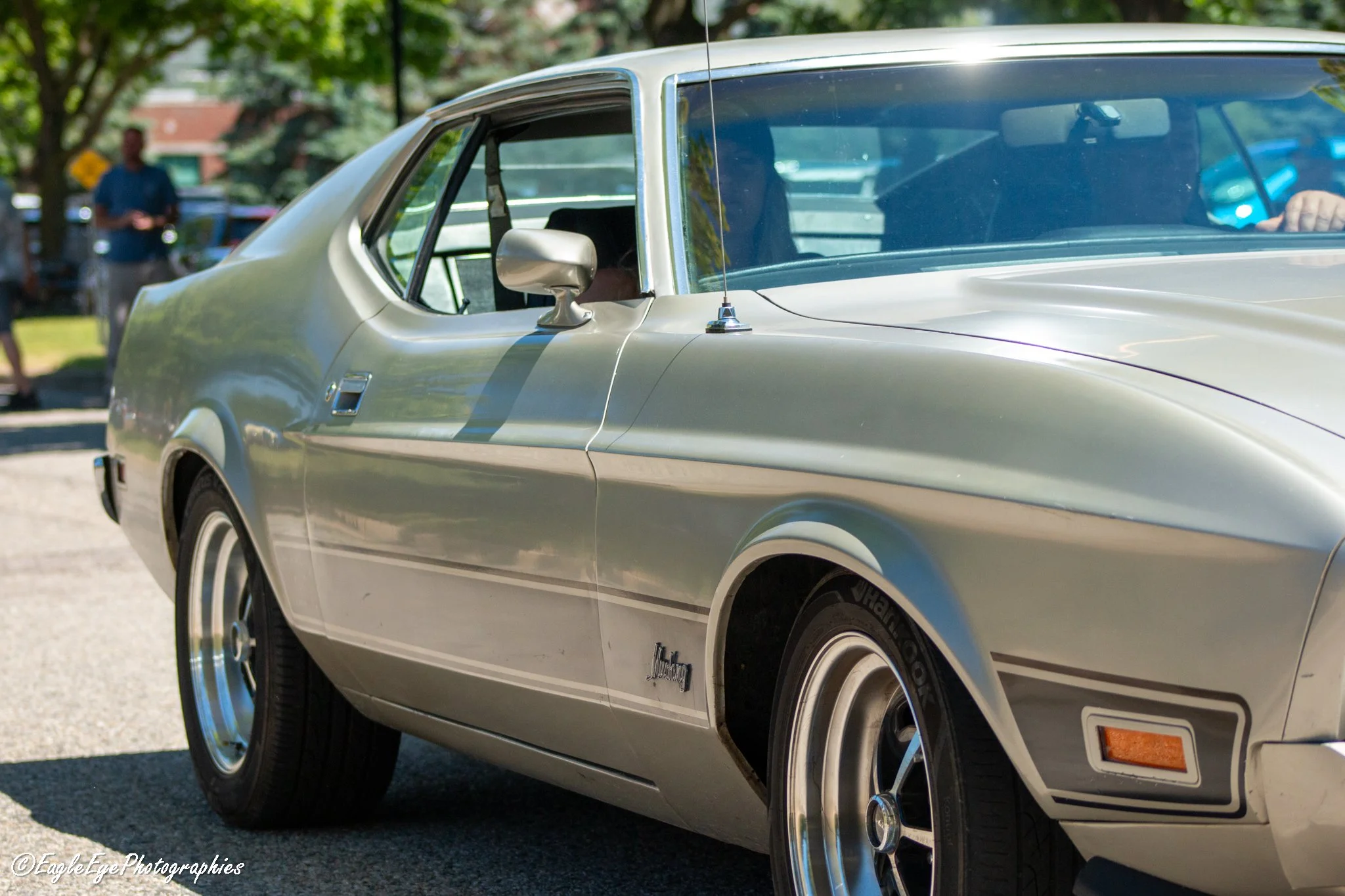Close-up view of a classic silver sports car parked outdoors, showing the front side, side mirror, wheel, and part of the windshield with a person and trees reflected in the glass.