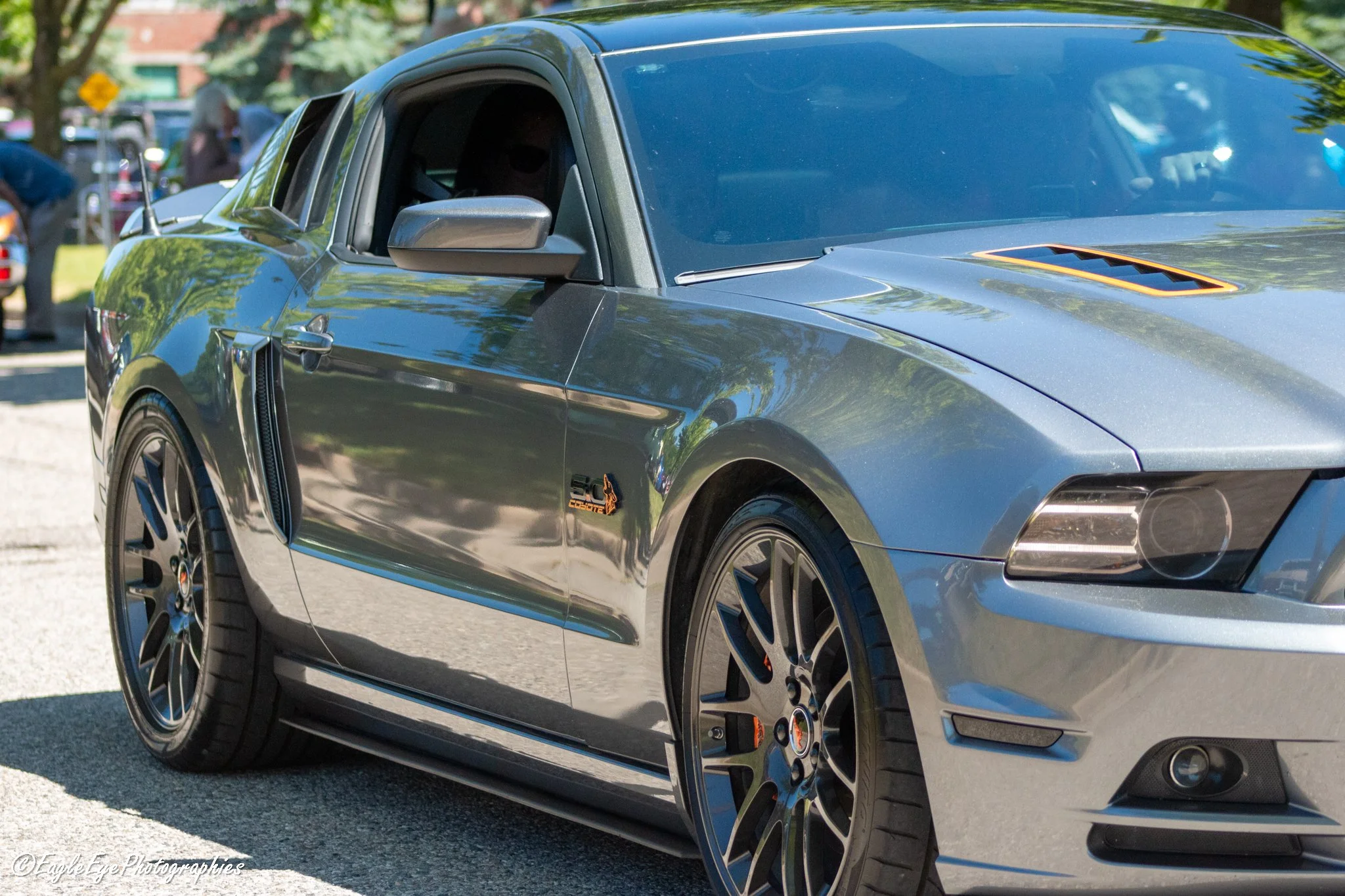 Close-up of a sleek black sports car parked outdoors with trees and people in the background.