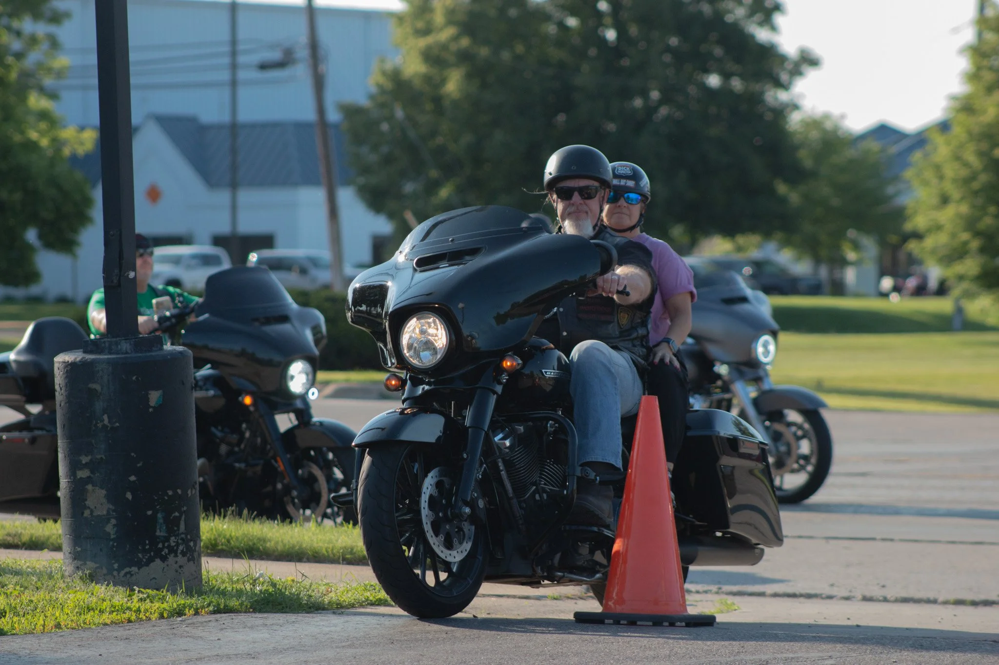 A man and woman on a motorcycle participating in a motorcycle safety course. The man is wearing sunglasses, a helmet, and a gray shirt. The woman is behind him, also wearing a helmet with blue reflection sunglasses. The motorcycle is black, and there