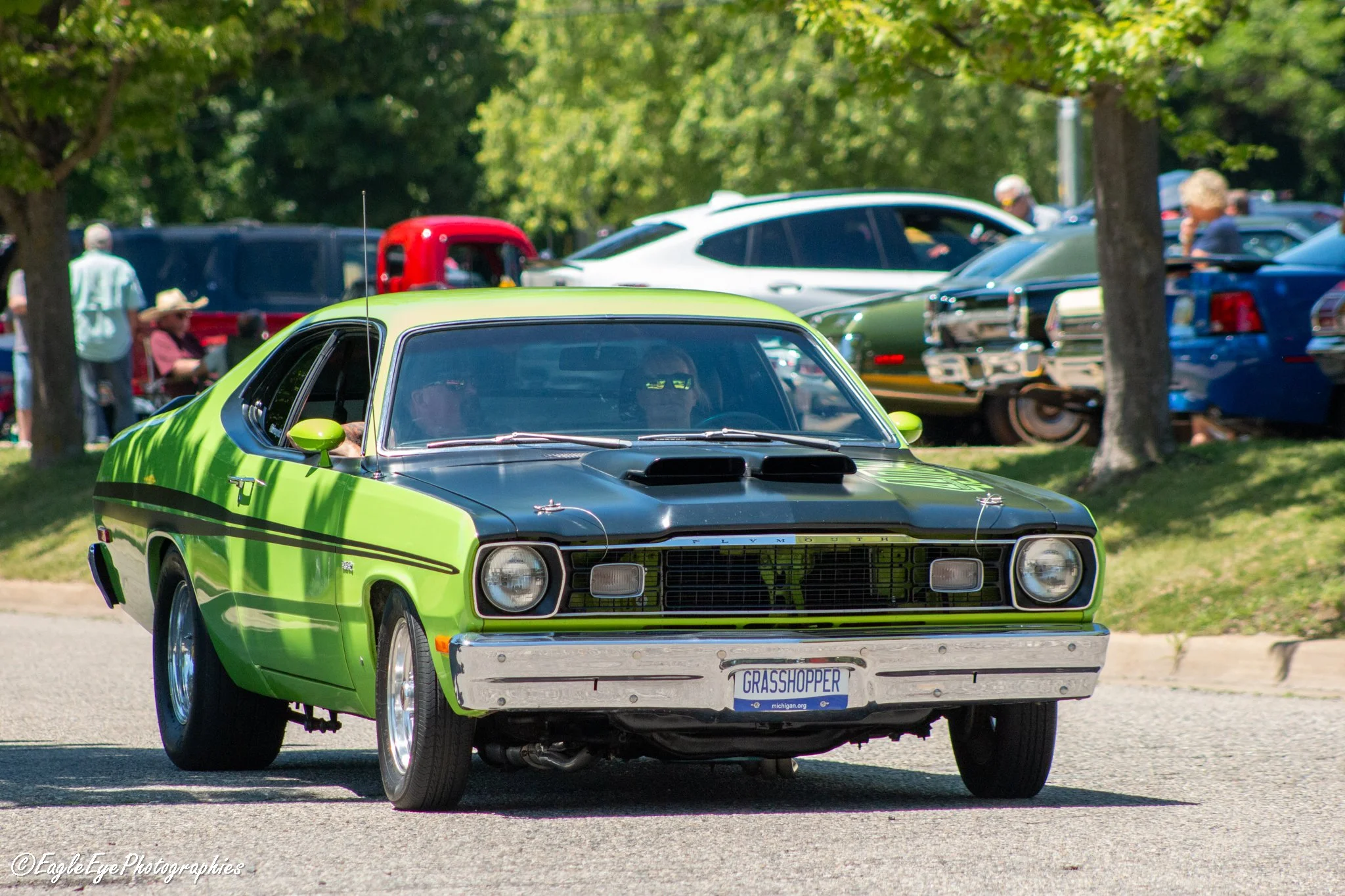 A bright green classic muscle car with a custom license plate that says 'GRASSHOPPER' parked outdoors during a car show, with other cars and people visible in the background.