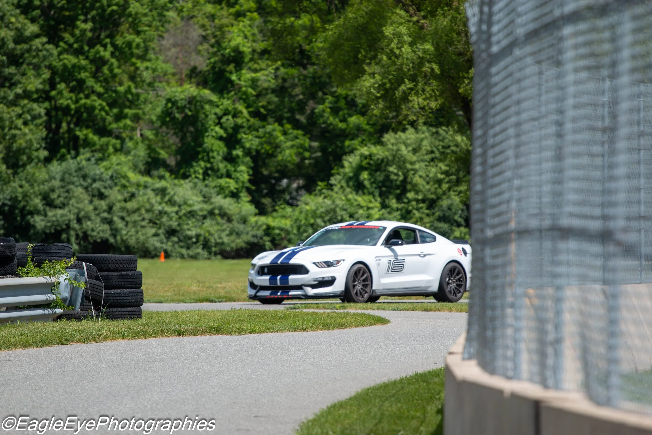 A white sports car with racing stripes on a racetrack, partially obscured by a barrier on the right and stacks of tires on the left, surrounded by green trees.