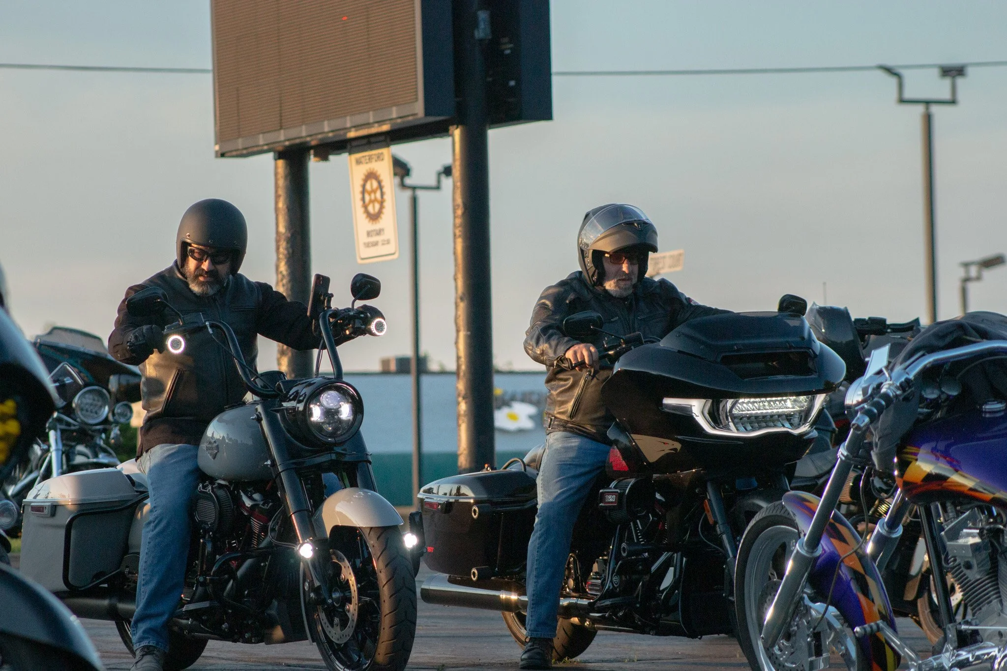 Two men riding motorcycles parked in a lot, both wearing helmets and leather jackets, with a large digital billboard and street signs in the background during dusk.
