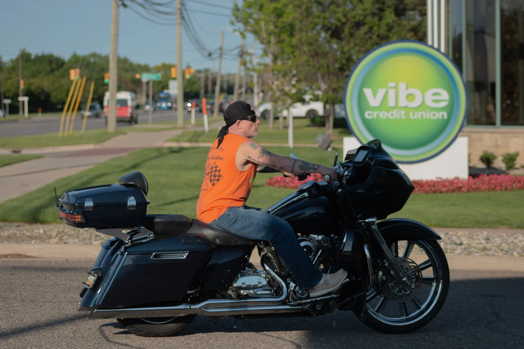 A man wearing sunglasses and an orange sleeveless shirt riding a black motorcycle on a city street, with a green Violet credit union sign in the background.