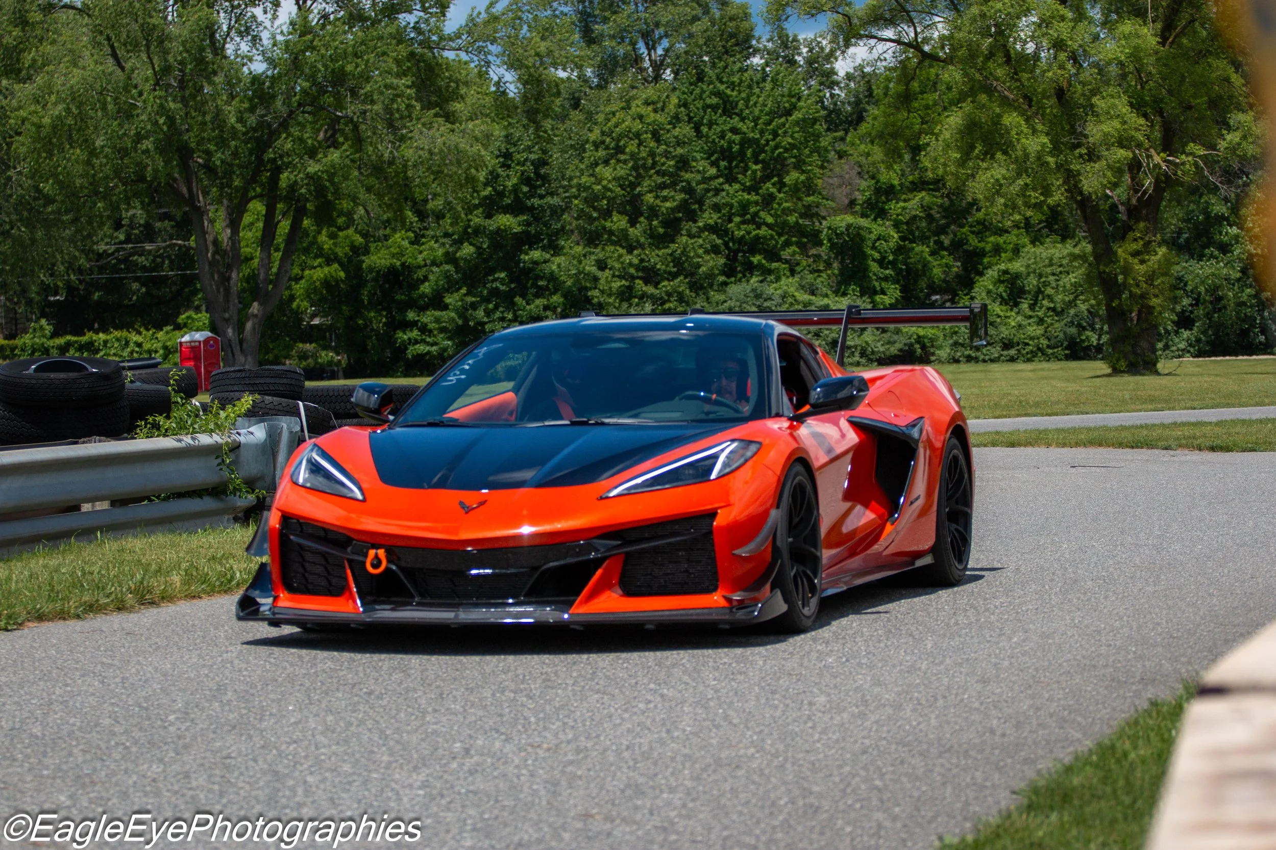 A red sports car with black accents driving on a road with a metal guardrail on the side, surrounded by lush green trees.