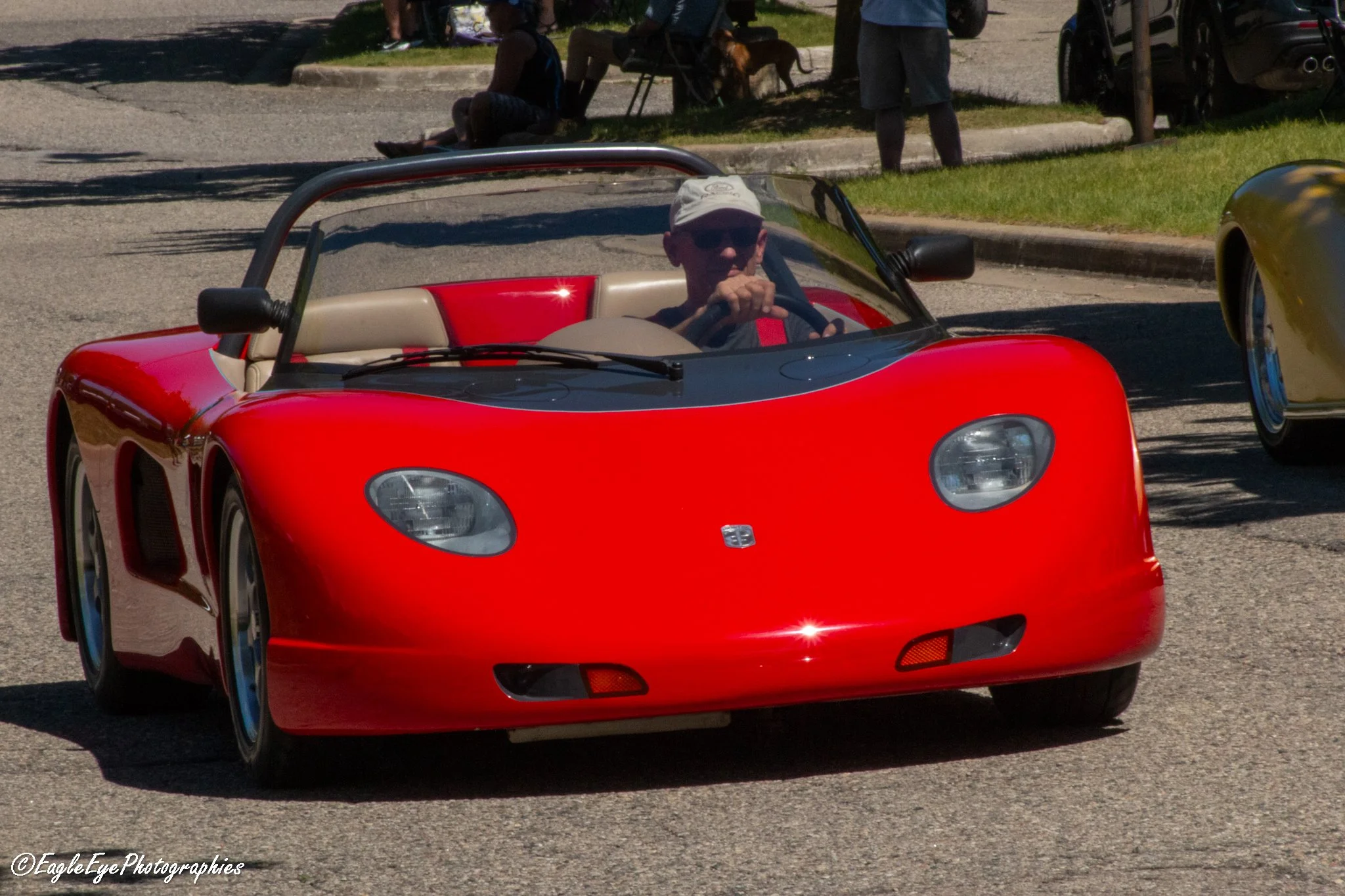 A man wearing sunglasses, a white cap, and a black shirt driving a red sports car with a beige interior on a sunny day.