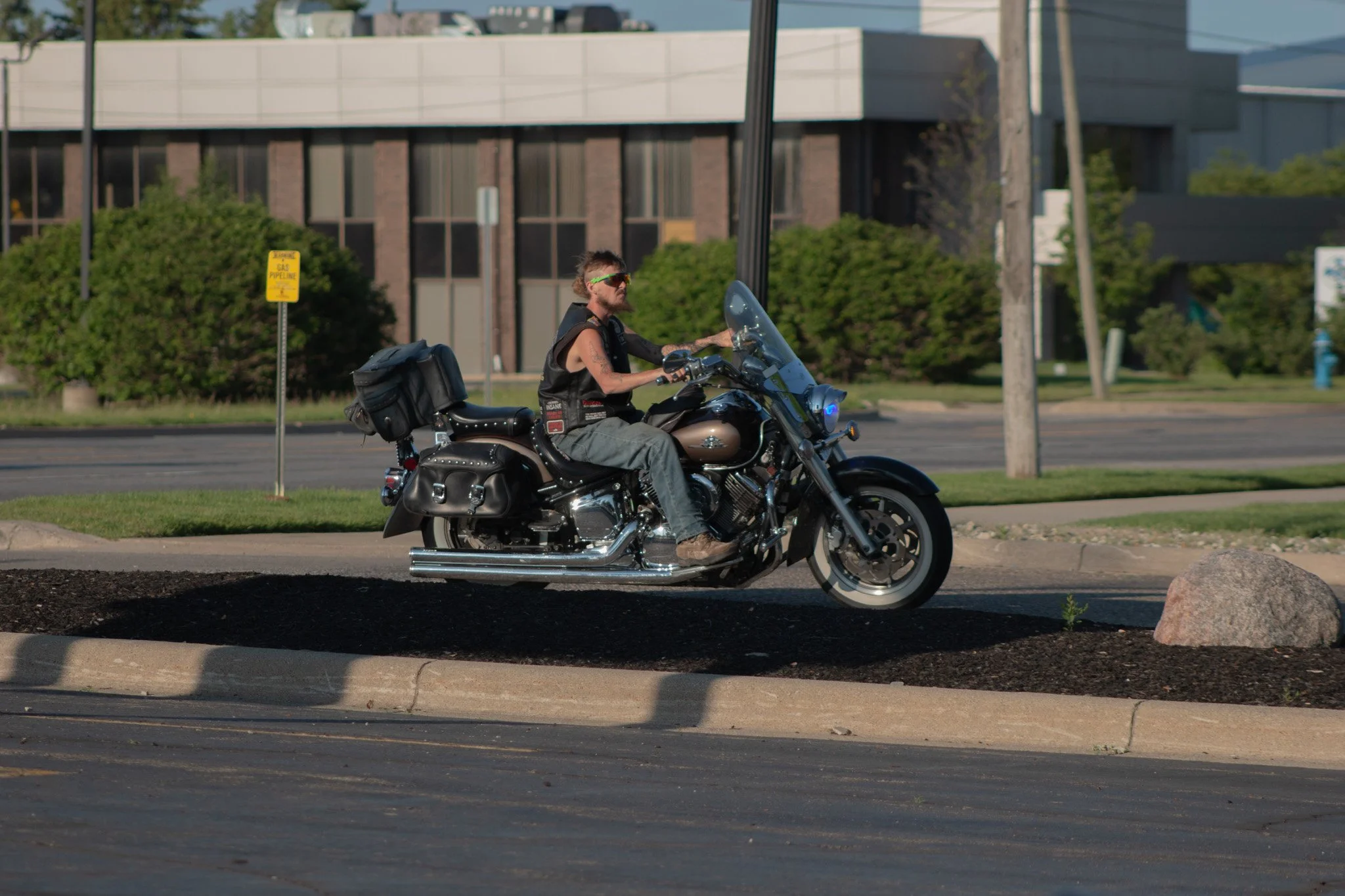 Man riding a black motorcycle on a city street with green trees, a building, utility pole, and a yellow gas pipeline sign in the background.
