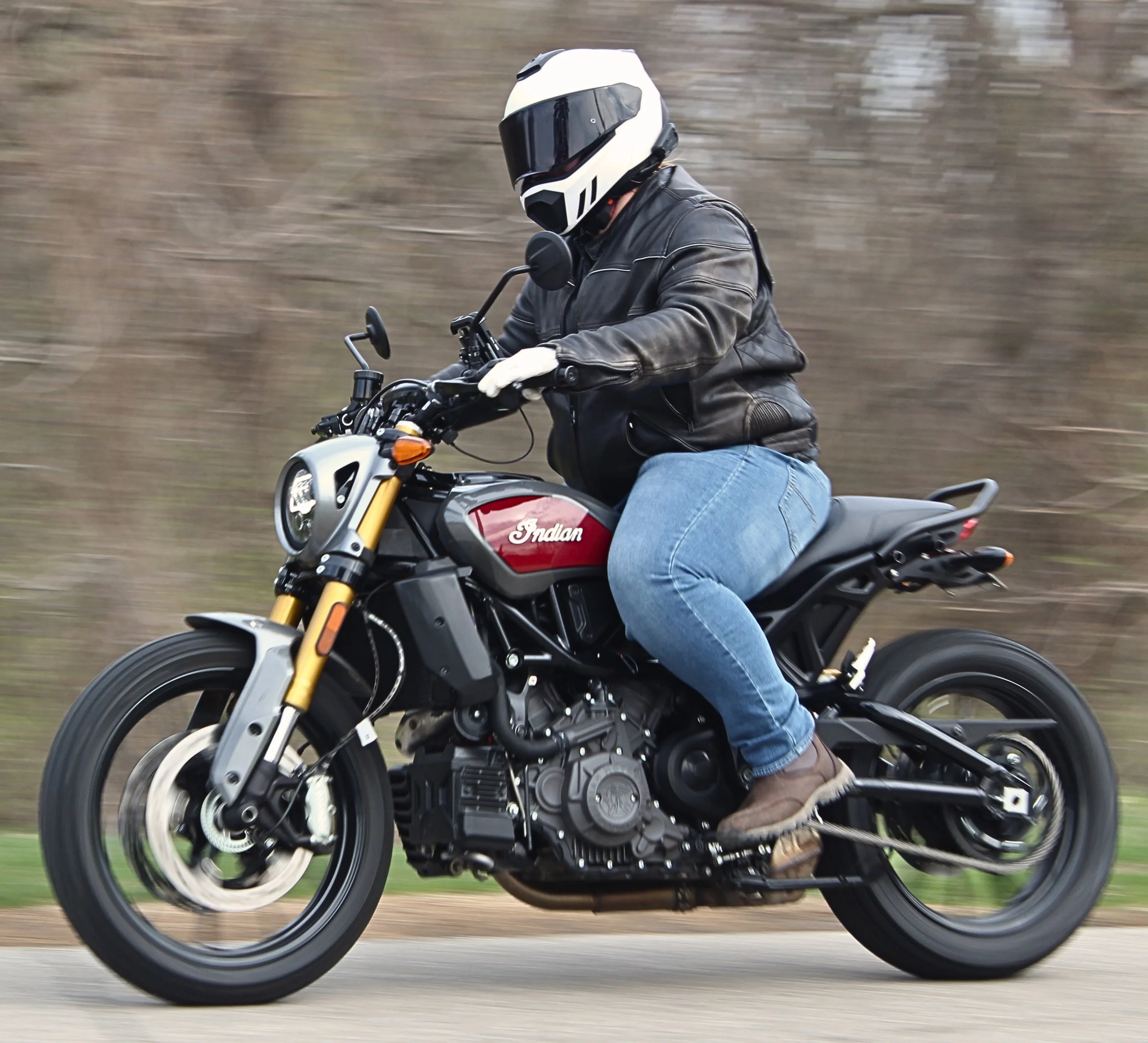 Person riding an Indian motorcycle wearing a white helmet, black leather jacket, and jeans on a blurred outdoor background.