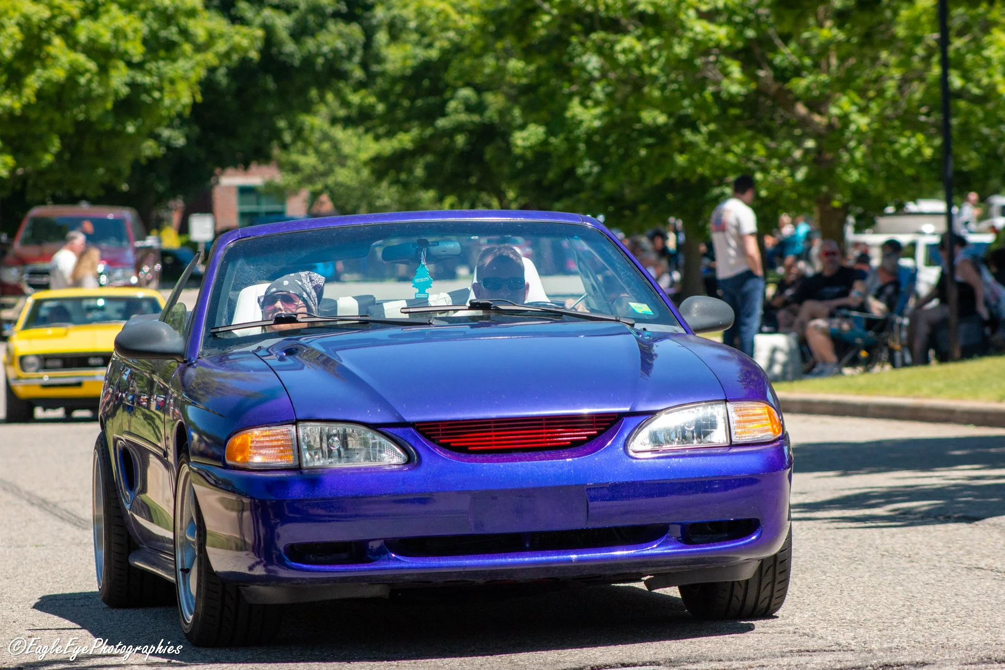 A shiny purple sports car driving in a park with trees and people in the background.