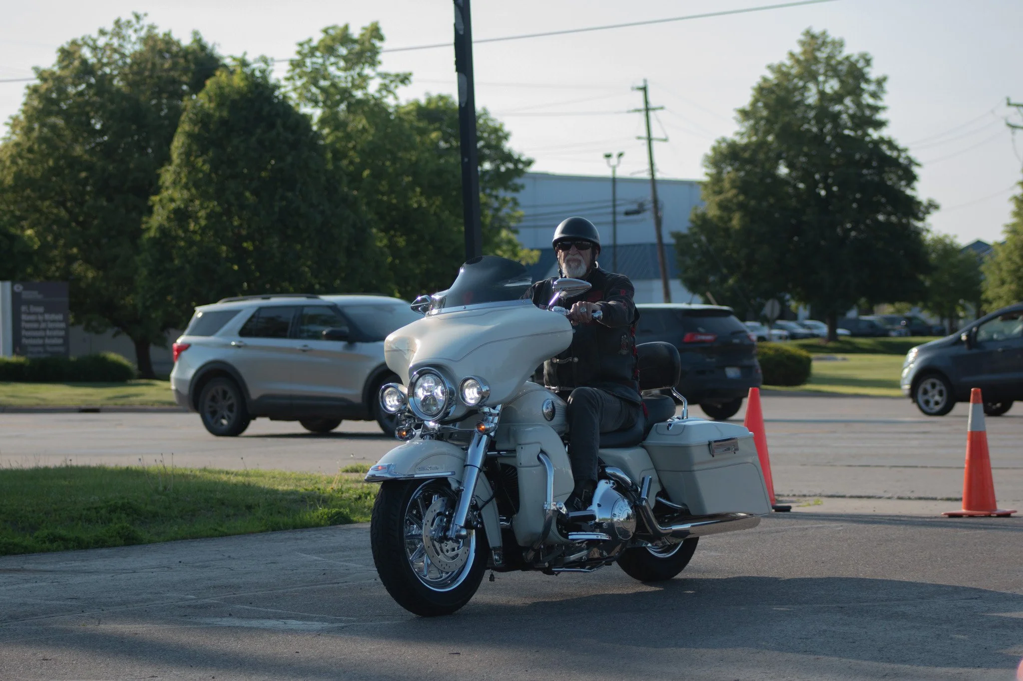 A man riding a white motorcycle with a large front fairing and windshield, wearing a black helmet and sunglasses, on a parking lot with orange traffic cones and other parked cars in the background.